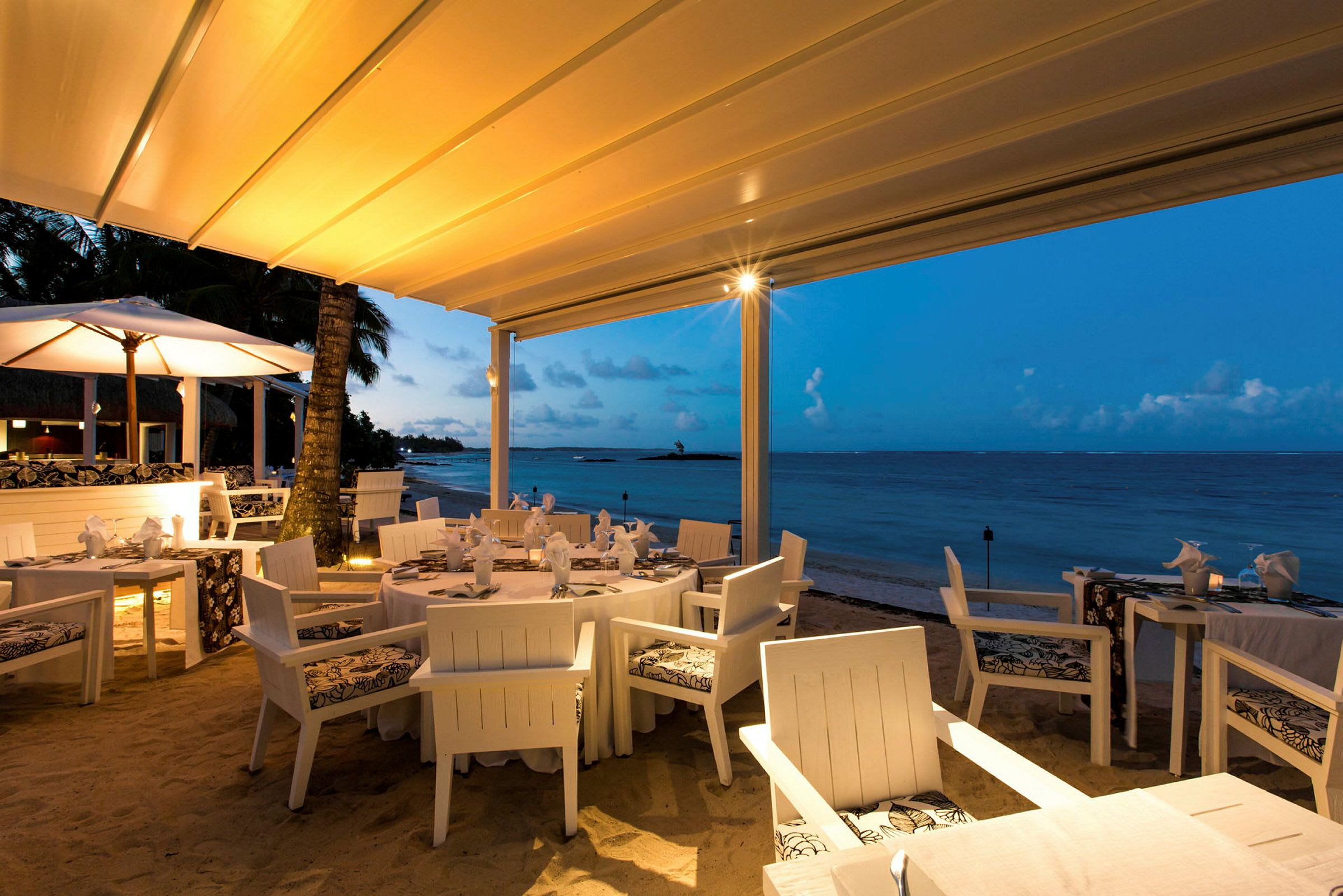 En restaurant med hvit møblering ligger ved stranden under en teltlignende takstruktur. Havet vises i bakgrunnen med en klar blå himmel og diskrete skyer. (AI-generert tekst)