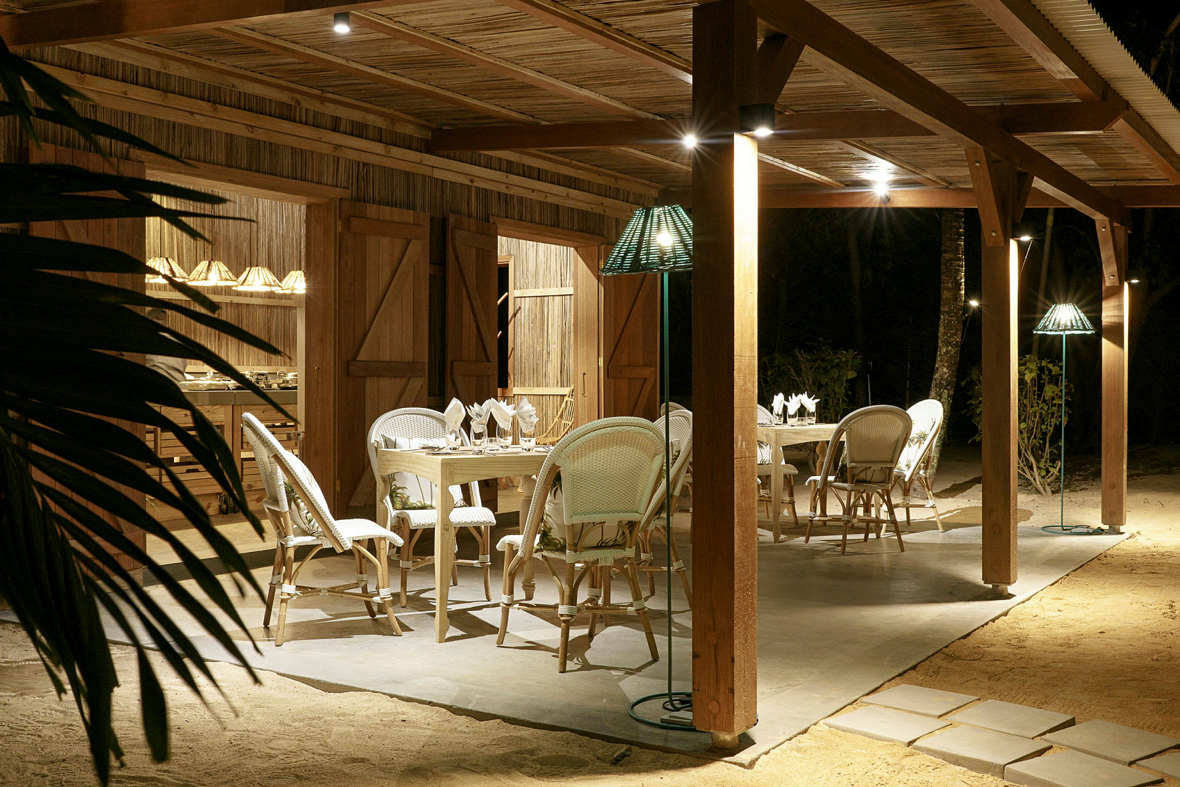 Utendørsområde med bord og stoler under en trepergola, kveldsbelysning og sandunderlag. (AI-generert tekst)