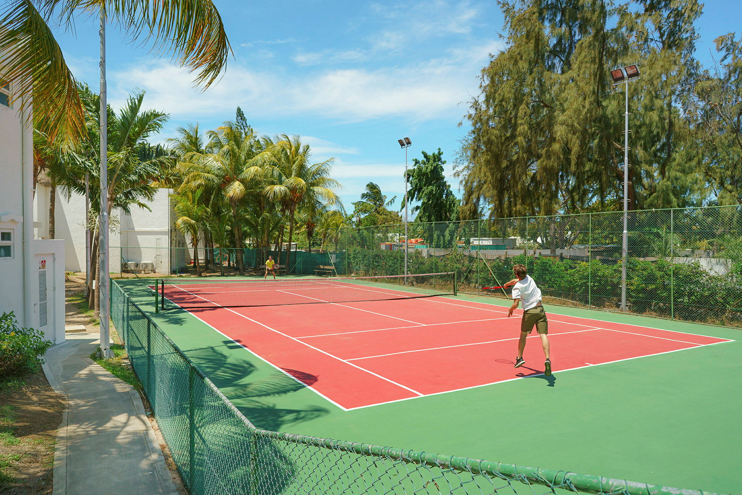 Utendørs tennisbane med to spillere nær et gjerde og palmetrær. (AI-generert tekst)