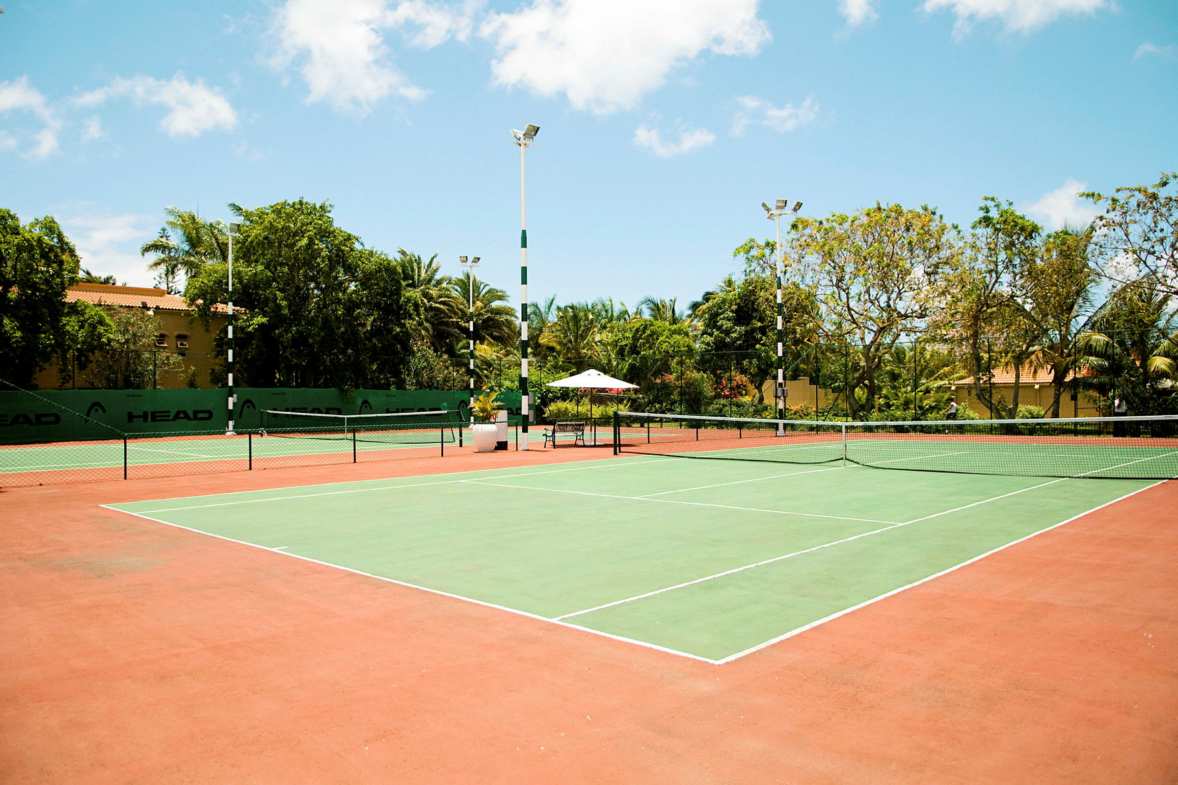 En tennisbane med grønt spilleområde og røde omkringliggende områder, omgitt av trær og bygninger. (AI-generert tekst)