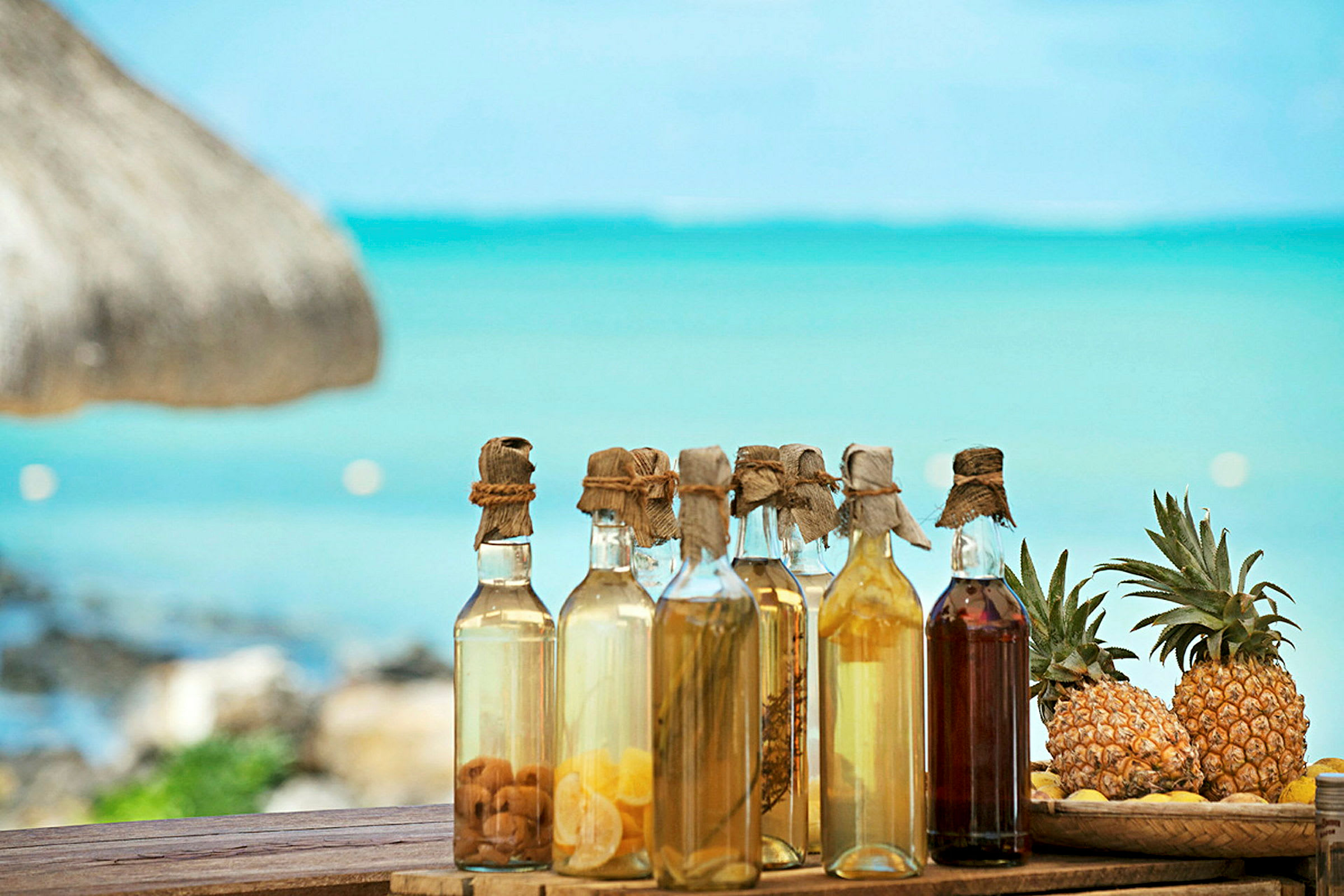 Flasker med forskjellige drikker og ananasfrukter ved en strand med utsikt over vann. (AI-generert tekst)