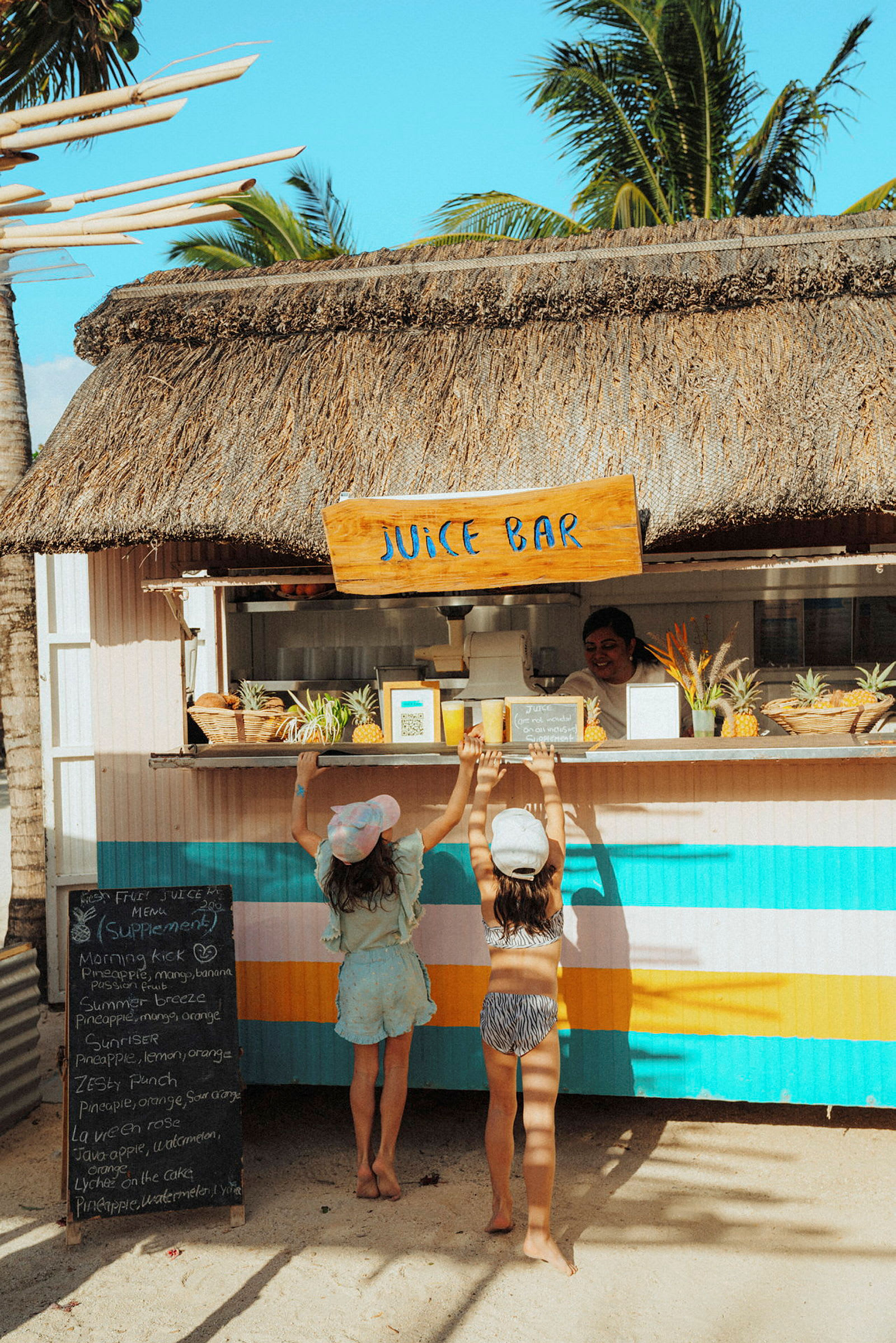 To personer står ved en juicebar med stråtak på et solrikt sted. (AI-generert tekst)