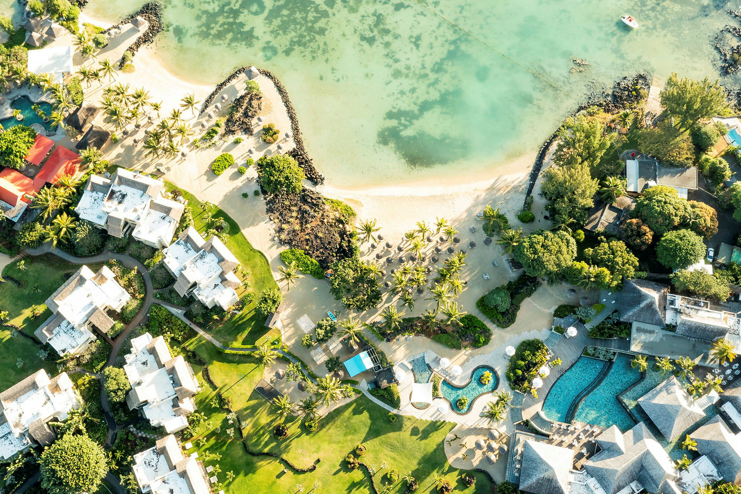 Flyfoto av hotellbygninger og bassengområder langs en sandstrand med palmer. (AI-generert tekst)
