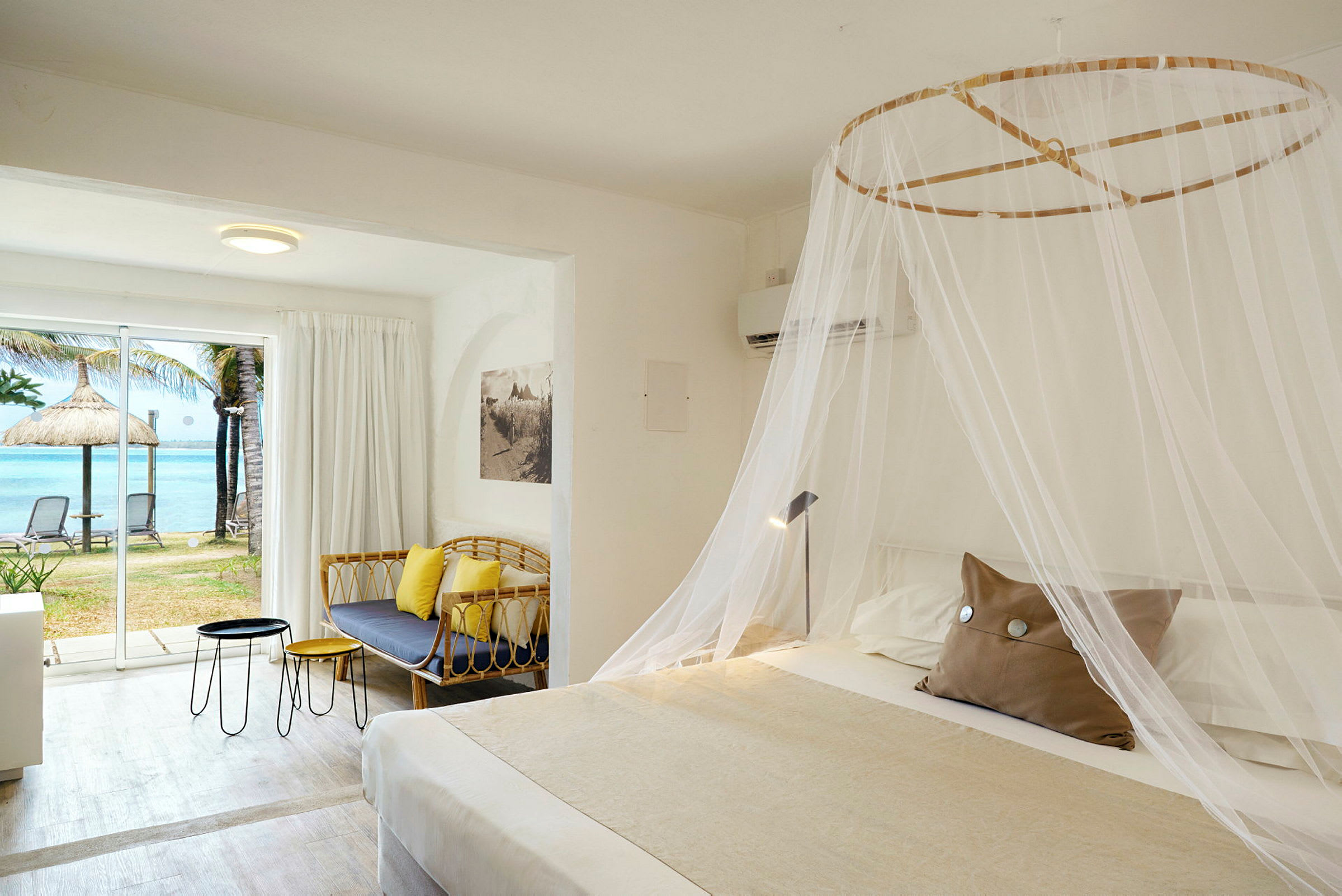 Hotellrom med en stor seng dekket med et myggnett, en sofa, bord og et stort vindu med utsikt over stranden og havet. (AI-generert tekst)