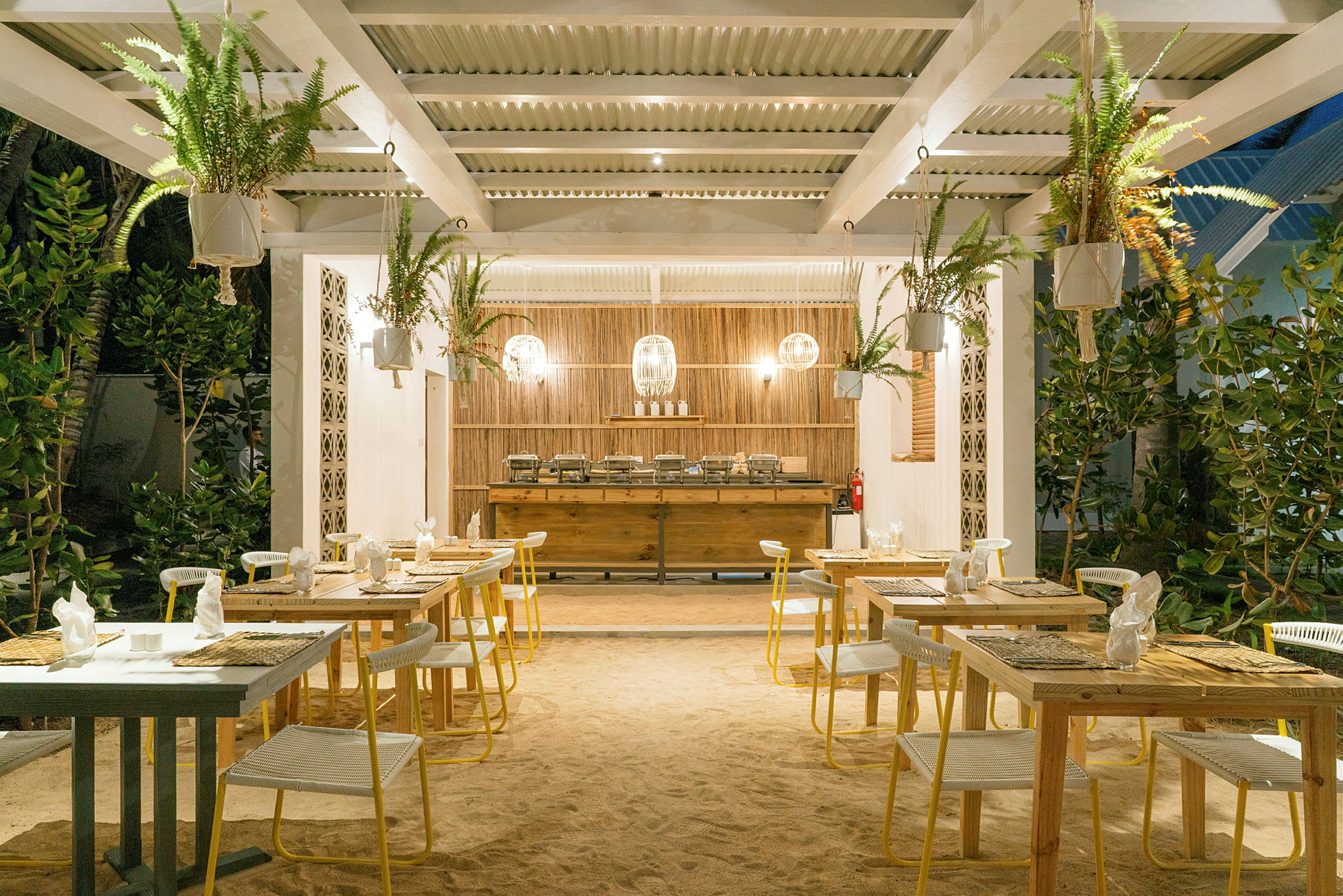 Utendørs restaurant med bord og stoler på sandgulv, omgitt av grønt. (AI-generert tekst)