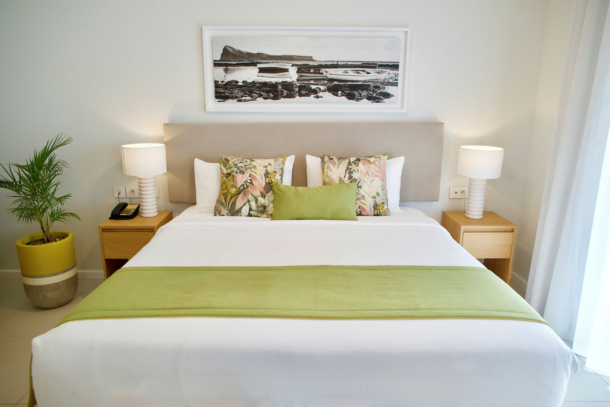 Et hotellrom med en stor dobbeltseng dekorert med puter i ulike farger og mønstre. På hver side av sengen står et nattbord med lampe, og en tavle henger over sengegavlen. En grønn plante i potte står ved siden av sengen. (AI-generert tekst)