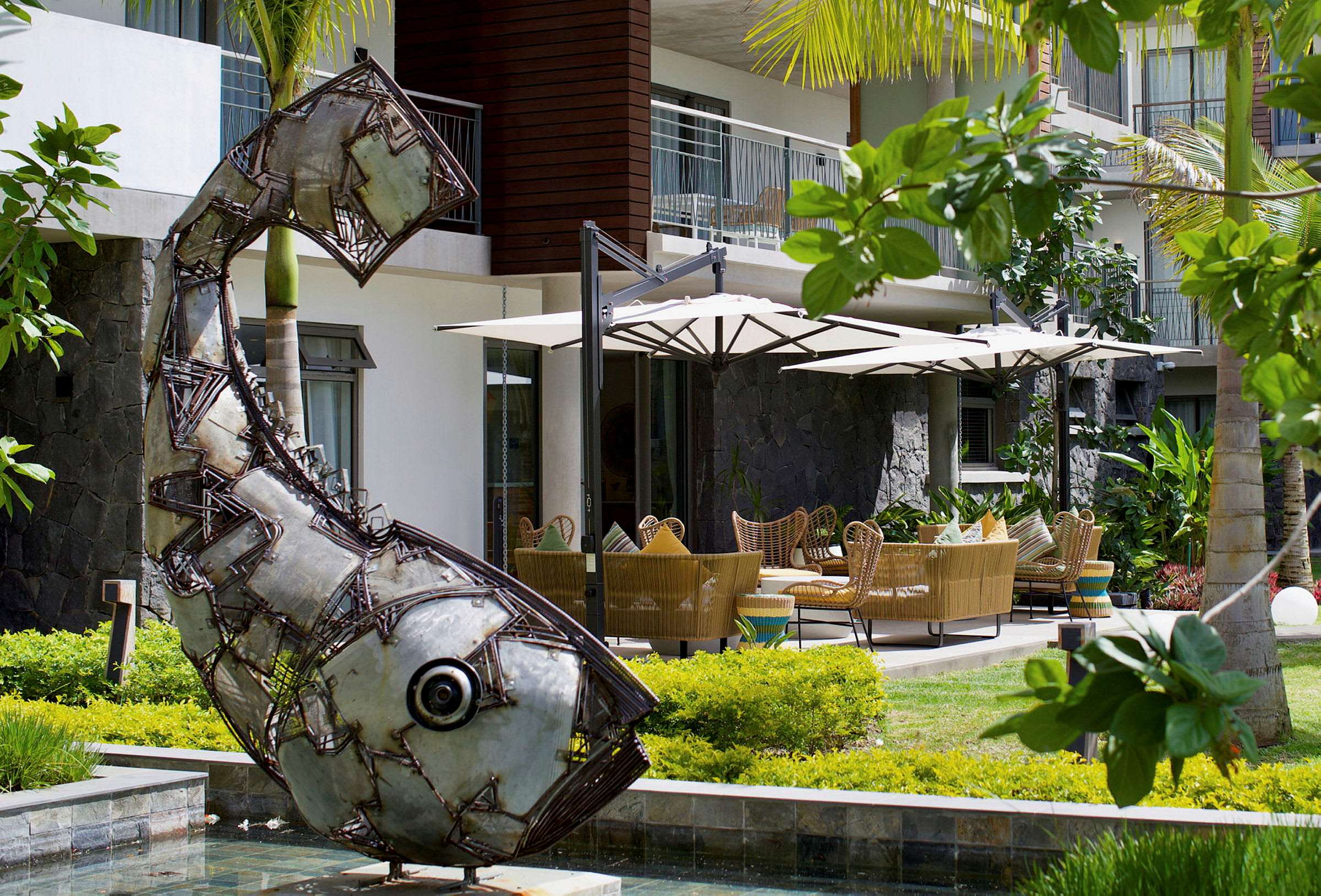 Metallskulptur formet som en fisk foran en terrasse med parasoller og sitteplasser. (AI-generert tekst)