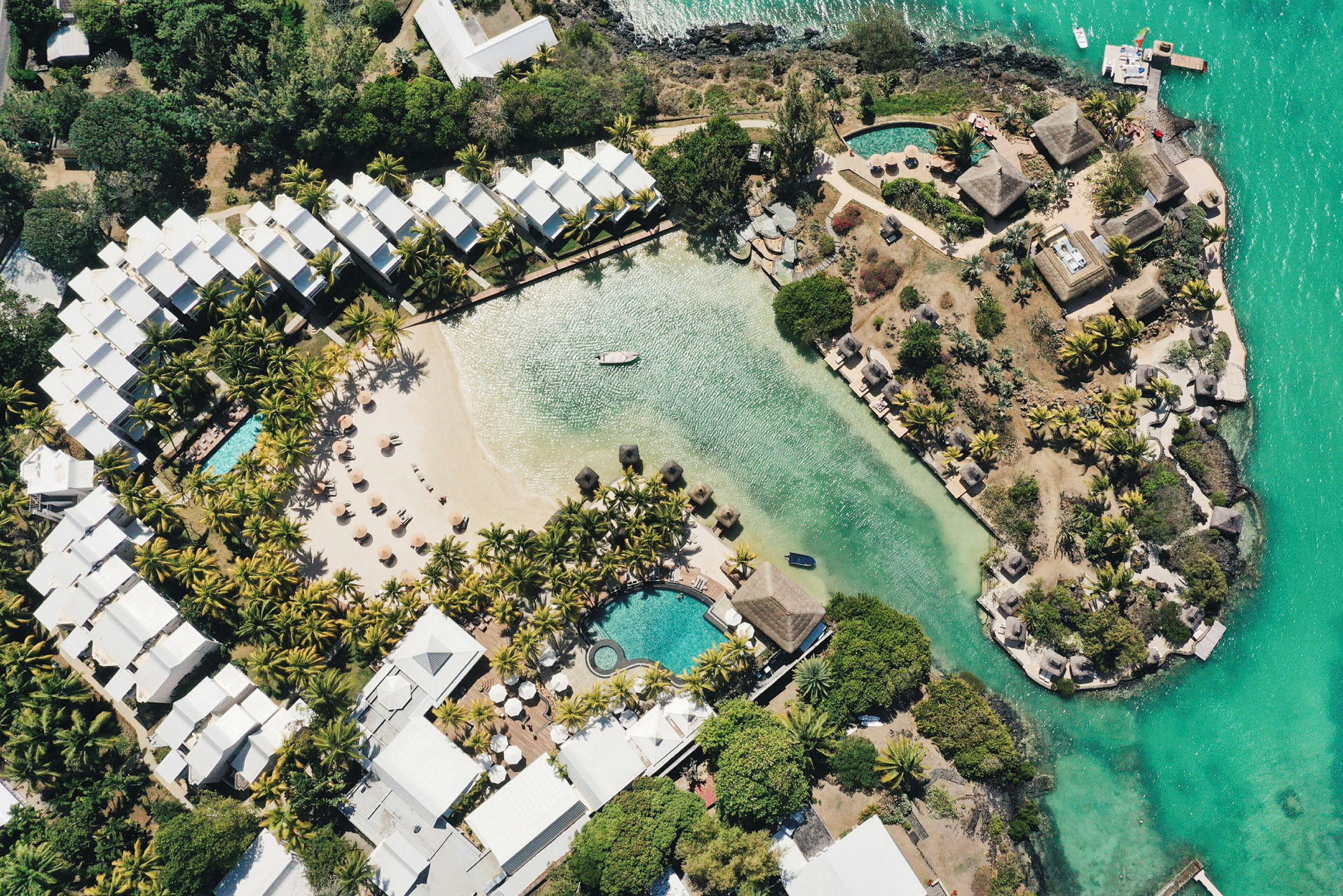 Flyfoto av ett hotellområde med basseng, en sandstrand og vegetasjon ved vannet. (AI-generert tekst)