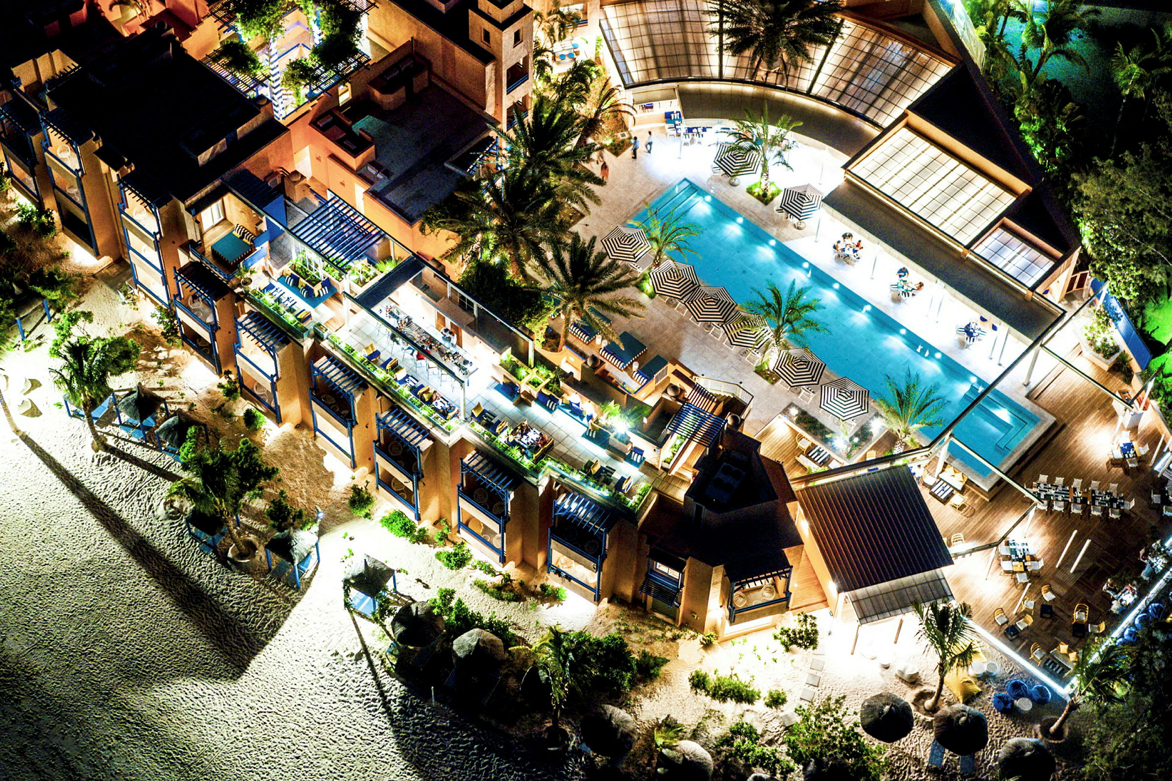 Fugleperspektiv over en resort med basseng, solsenger, parasoller og omkringliggende bygninger. (AI-generert tekst)