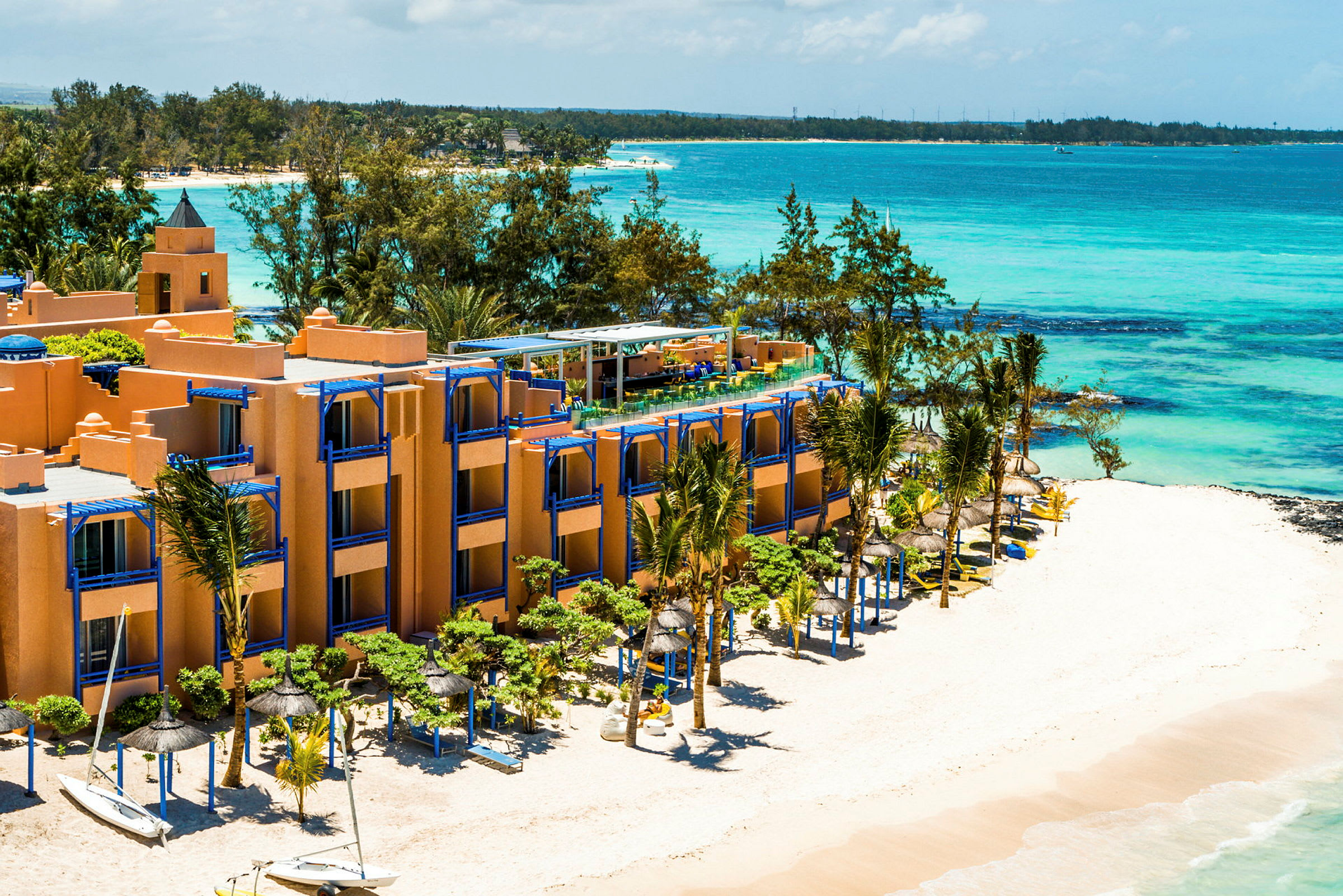 Hotellbygning ved en hvit sandstrand med blå parasoller og havet i bakgrunnen. (AI-generert tekst)