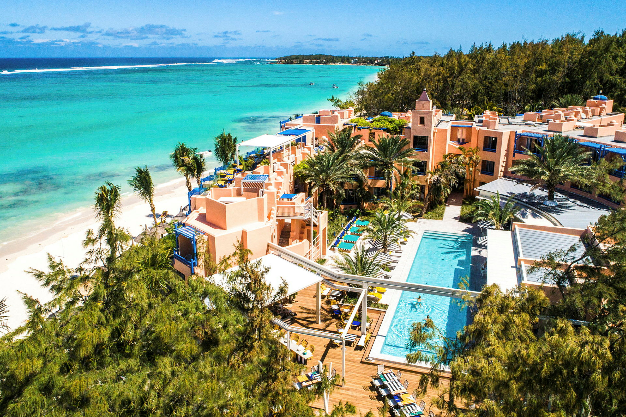 Flyvning over et hotellkompleks ved en tropisk strand med basseng og omgivende vegetasjon. (AI-generert tekst)
