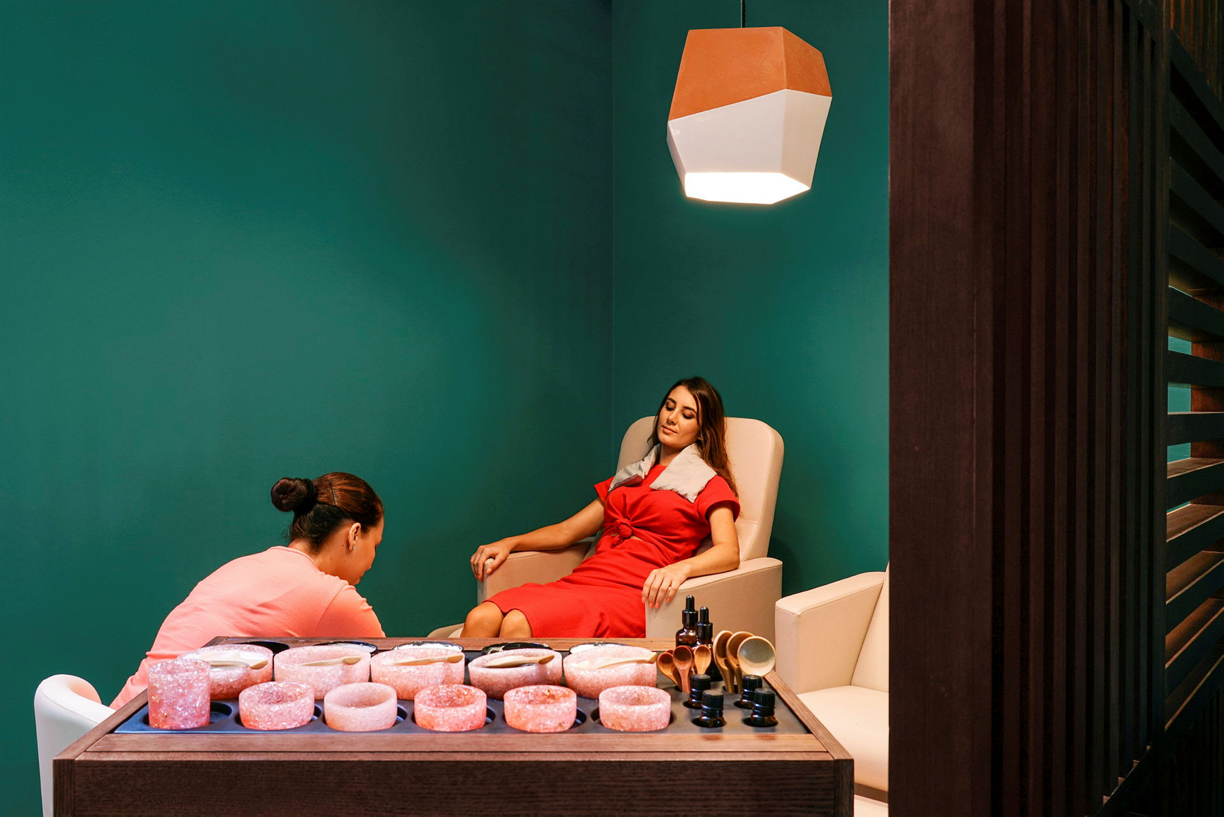 Et spa-rom med en person som får en behandling, mens en annen person assisterer. I forgrunnen er en trebenk med rosa saltsteiner og skjønnhetsprodukter. (AI-generert tekst)