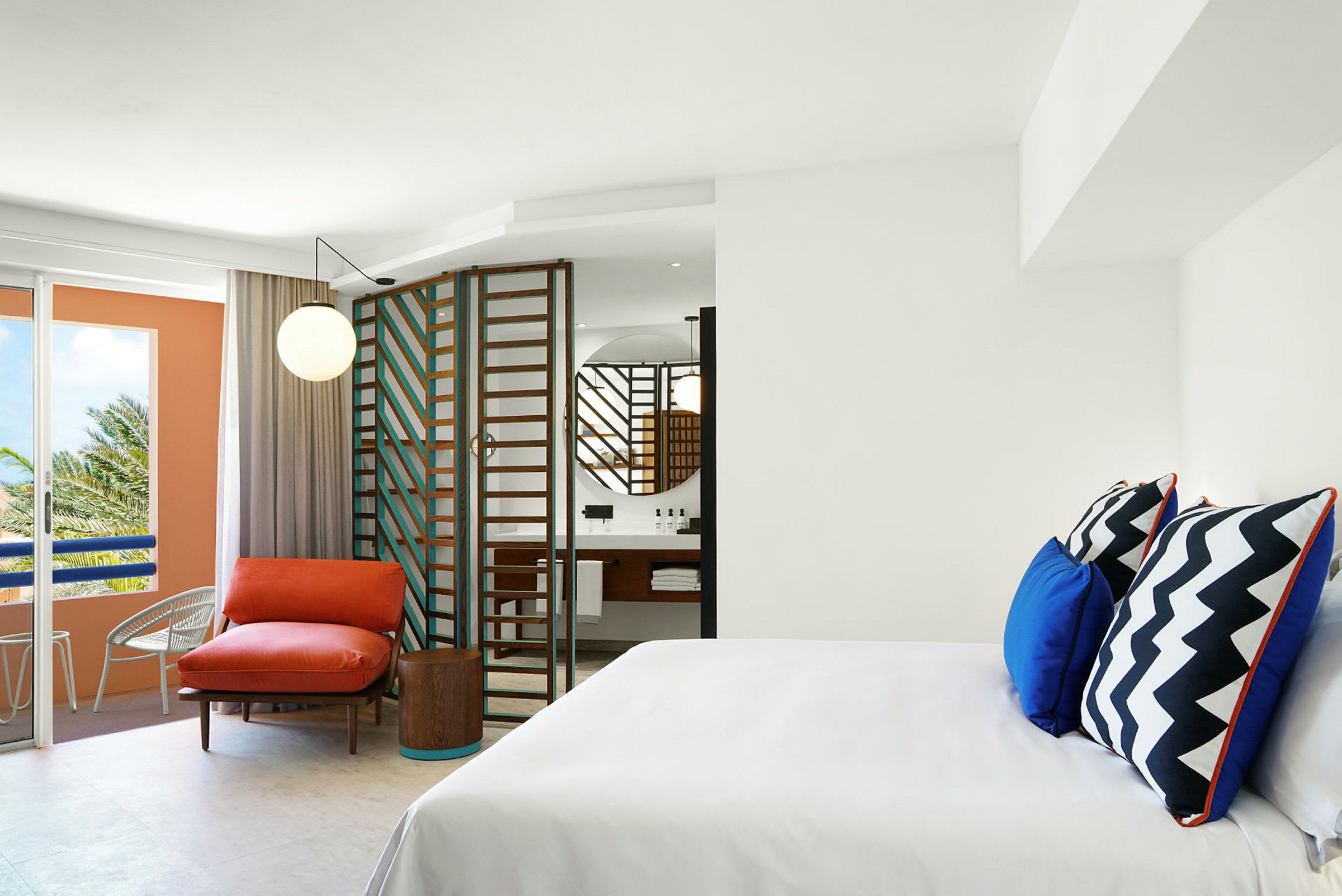 Et moderne hotellrom med hvite vegger, en fargerik lenestol og puter samt et bad med speil og en glassdør. (AI-generert tekst)