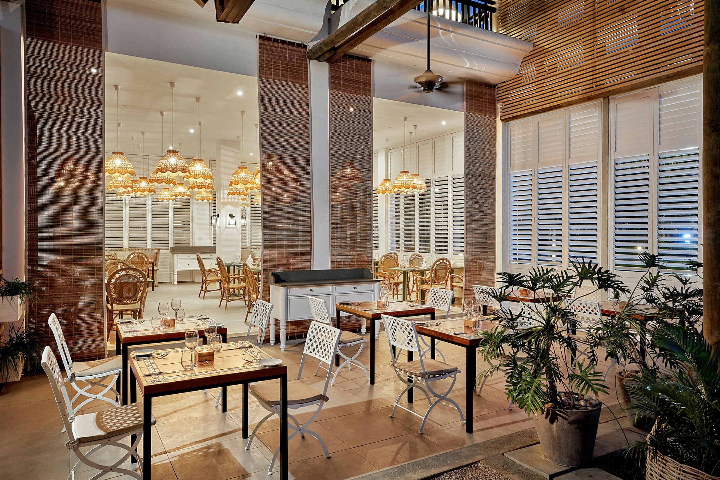 Innredning av en restaurant med bord og stoler i forskjellige seksjoner, omgitt av planter og med belysning fra designlamper. (AI-generert tekst)