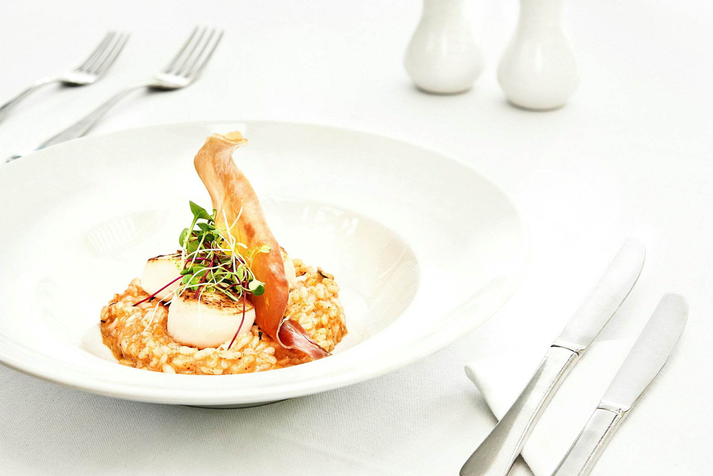 Tallerken med risotto, grillede kamskjell og en dekorasjon av mikrogrønt samt tynne skiver ingredienser. (AI-generert tekst)