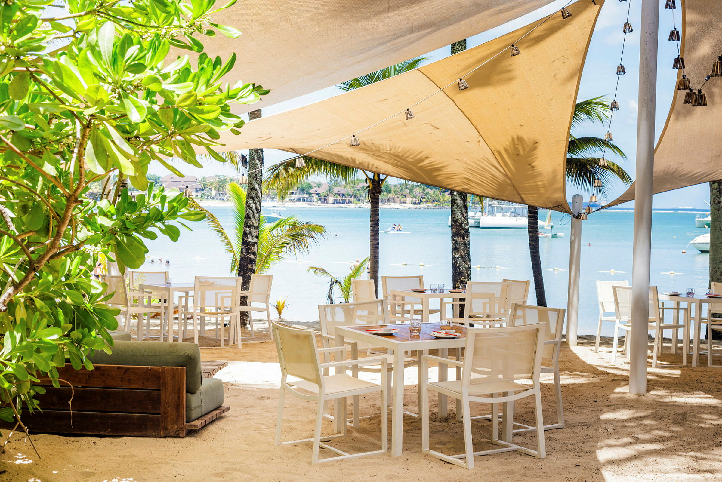 Utendørs restaurant med bord og stoler på sandstrand, omgitt av grønt og palmer med utsikt over havet. (AI-generert tekst)