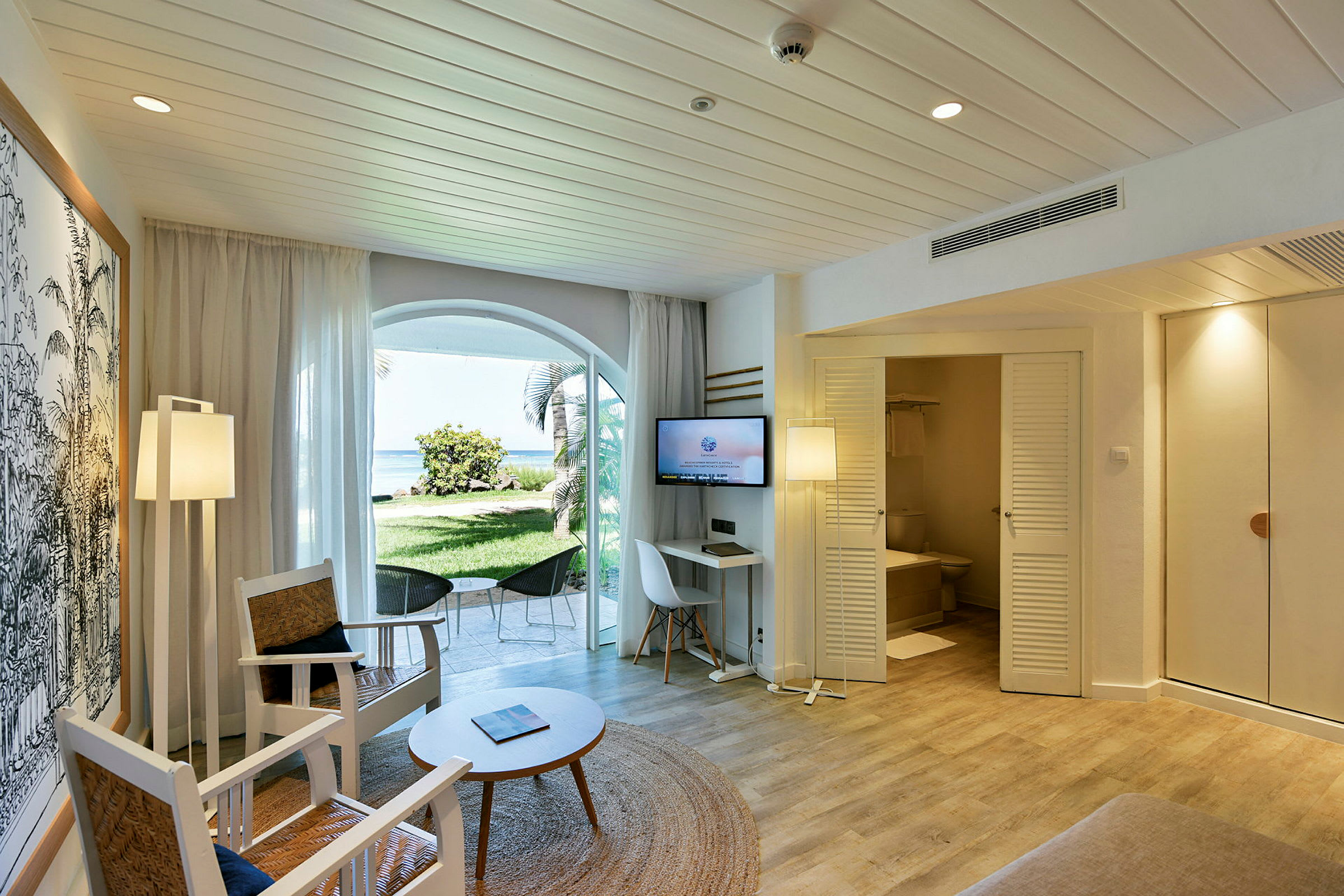 Et hotellrom med sittemøbler, bord og TV. En dør fører til et bad, og det er vinduer som vender mot en grønn hage og stranden. (AI-generert tekst)