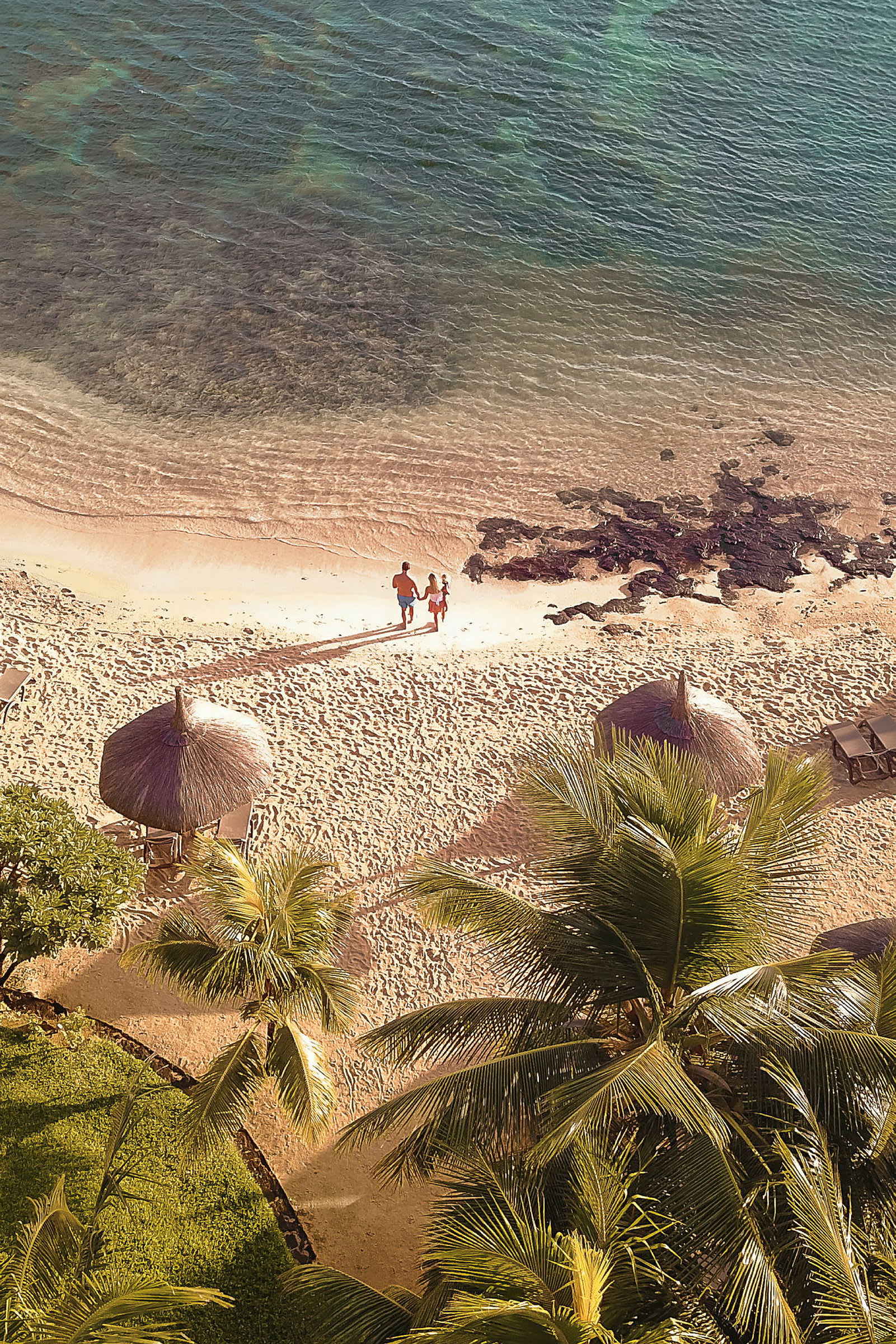To personer går på en sandstrand nær havet, omgitt av palmer og parasoller. (AI-generert tekst)