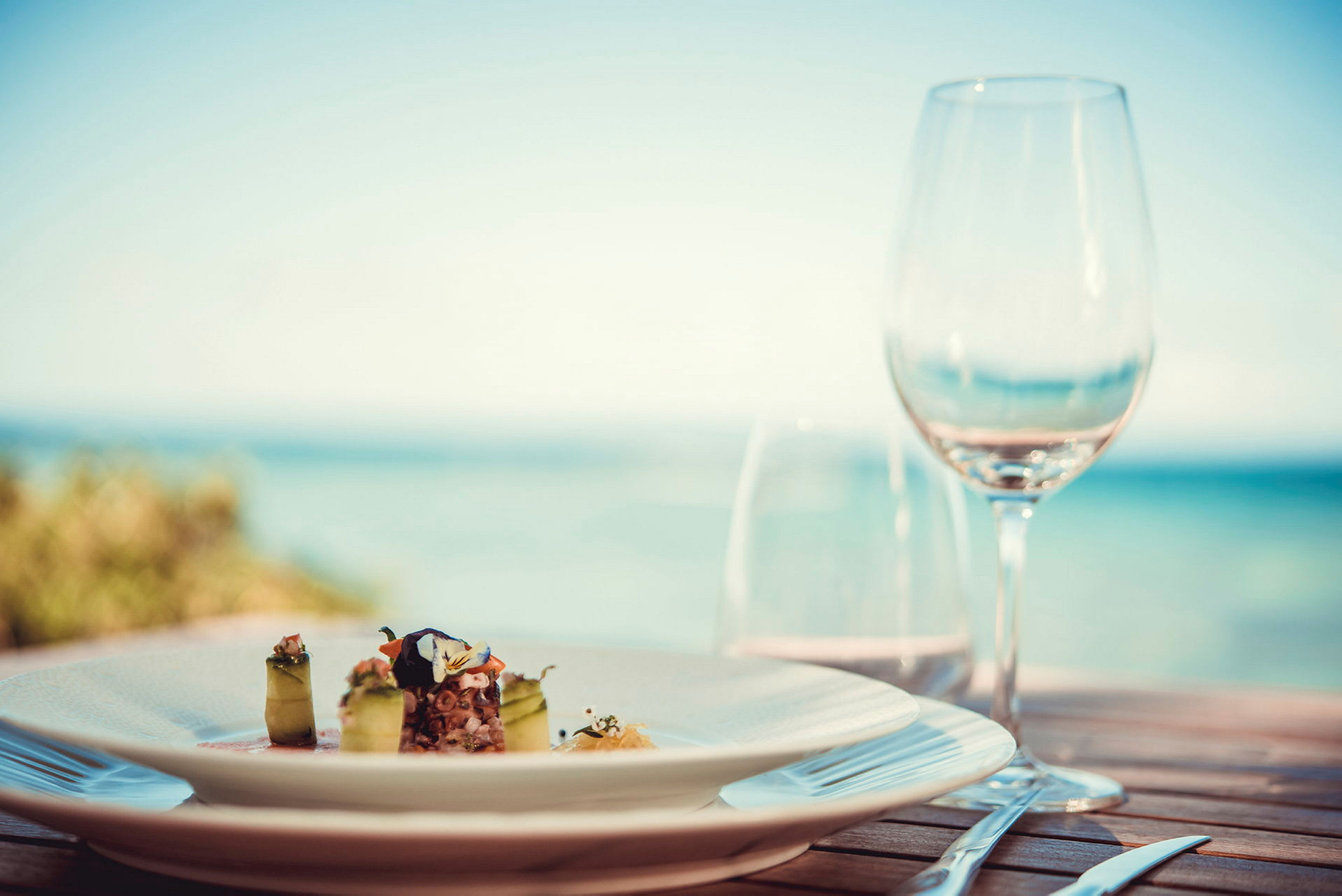 En tallerken med en elegant rett og et glass på et bord med havutsikt. (AI-generert tekst)