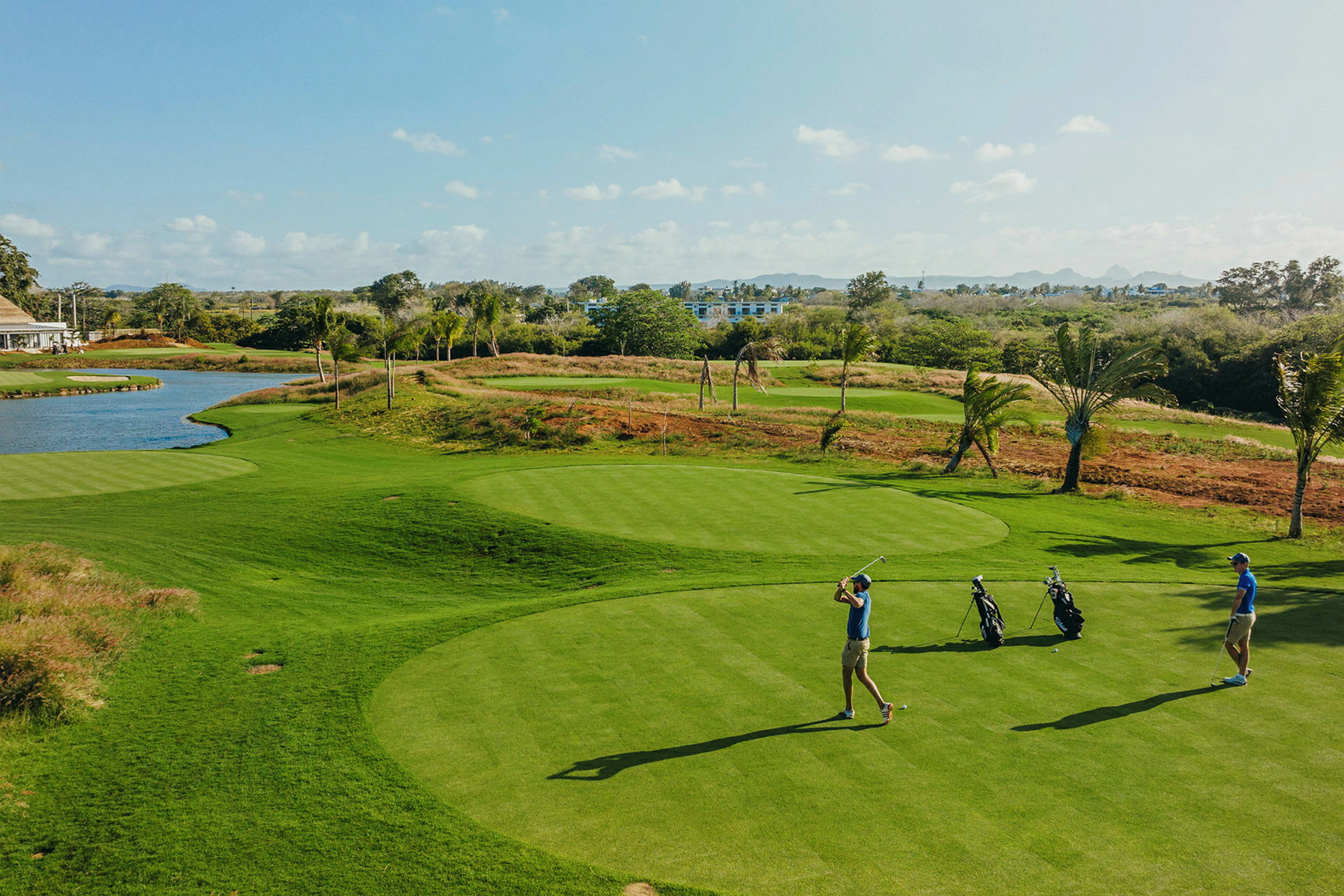 To personer spiller golf på en grønn golfbane med en liten innsjø og trær i bakgrunnen. (AI-generert tekst)