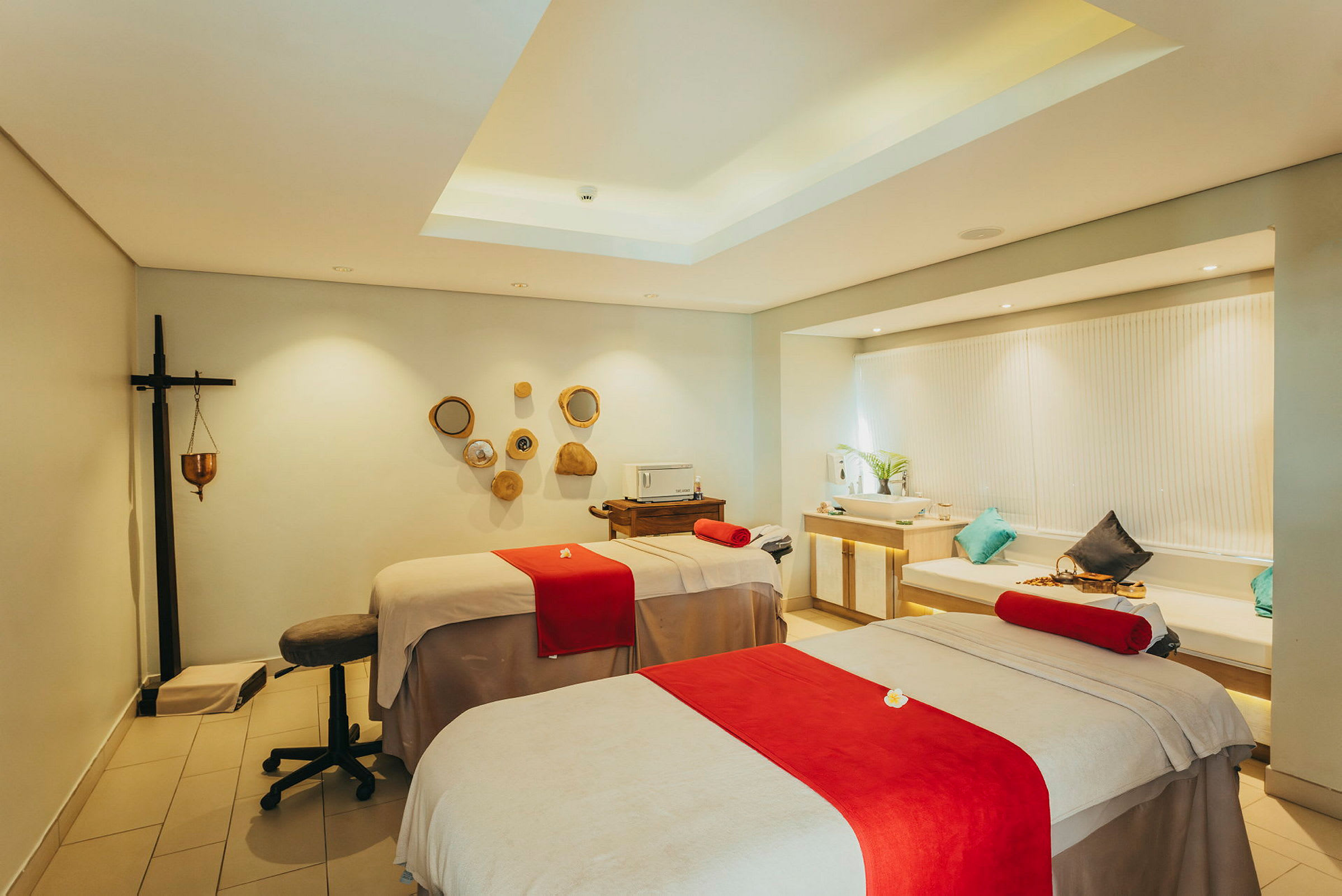 Et spa-rom med to massasjebord dekket med beige sengetøy og røde detaljer, innredet med dekorative elementer og moderne belysning. (AI-generert tekst)