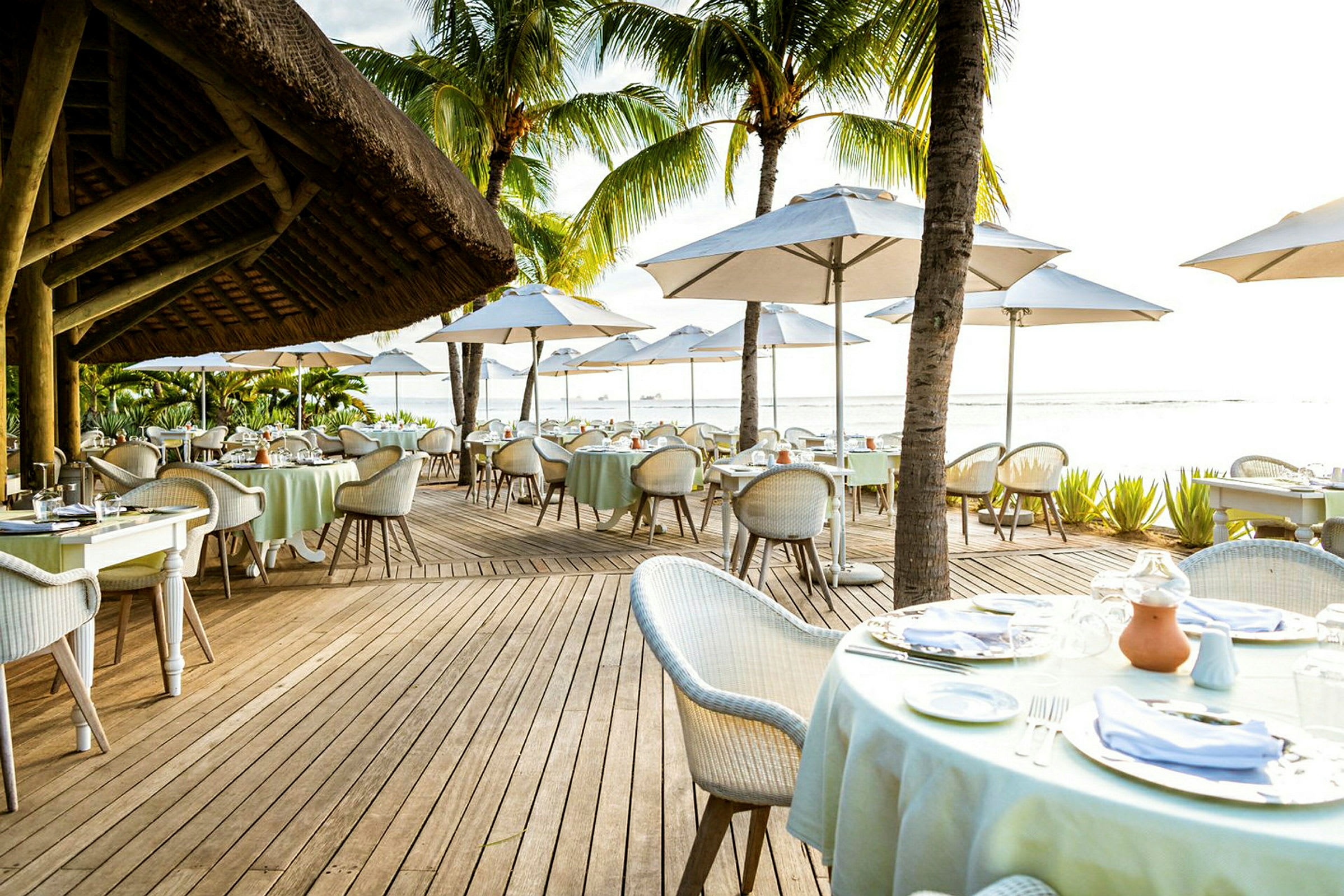Et sett med bord og stoler arrangert på en utendørsrestaurant med parasoller, plassert nær stranden. (AI-generert tekst)