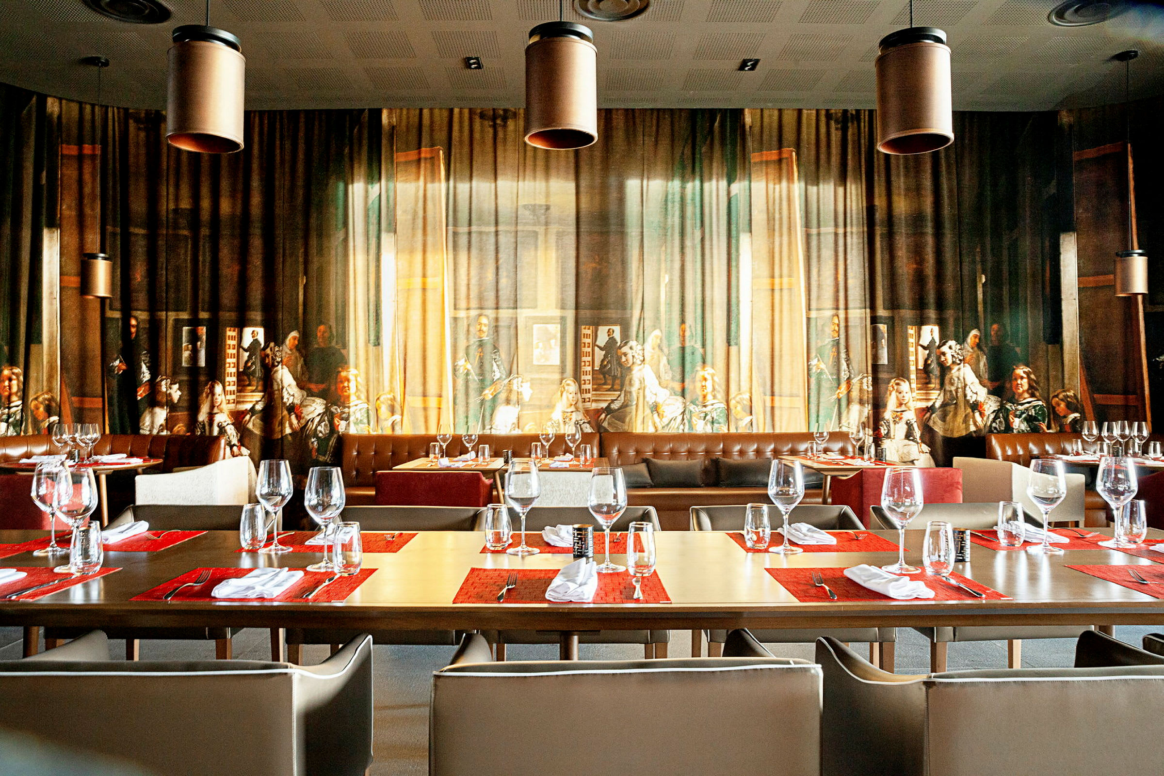 En restaurant med bord dekket med glass og servietter foran en vegg med et stort, detaljert kunstverk. (AI-generert tekst)