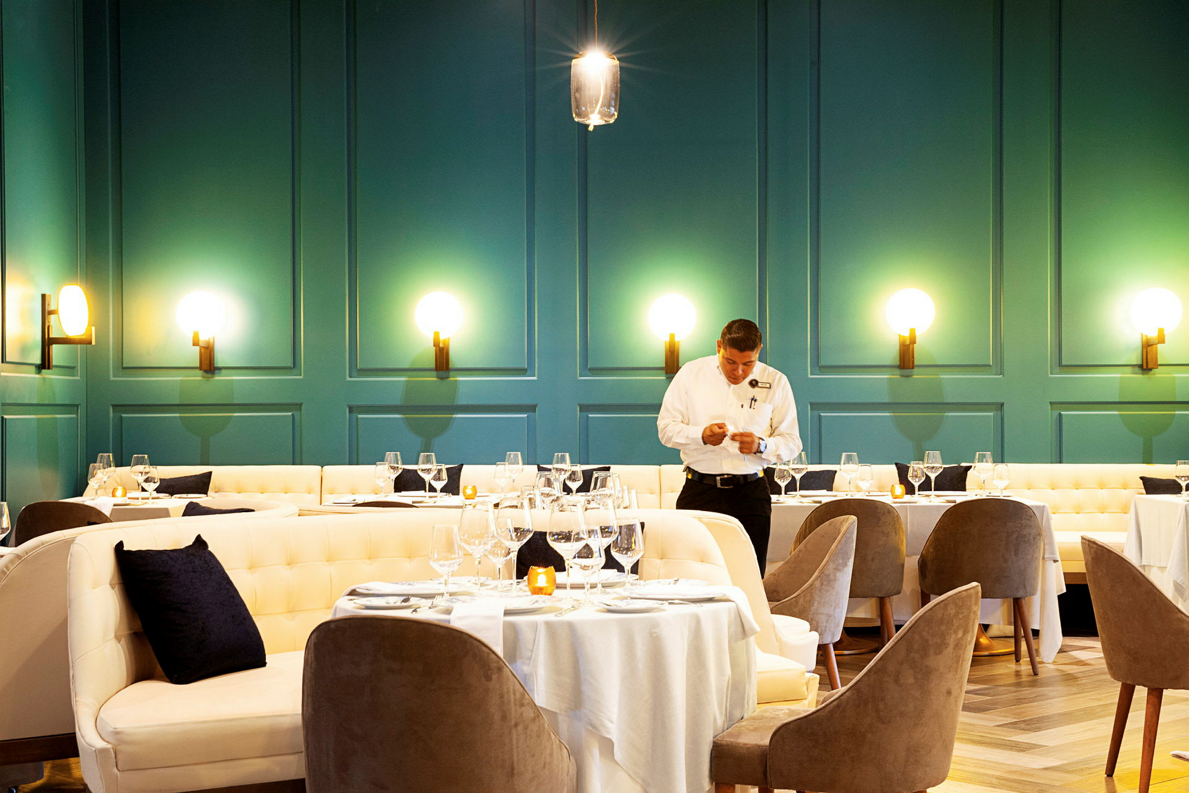 En elegant serveringslokale med dekkede spisebord omgitt av polstrede stoler og sofaer. Veggene er malt i en dyp grønn farge og dekorert med moderne lamper. (AI-generert tekst)