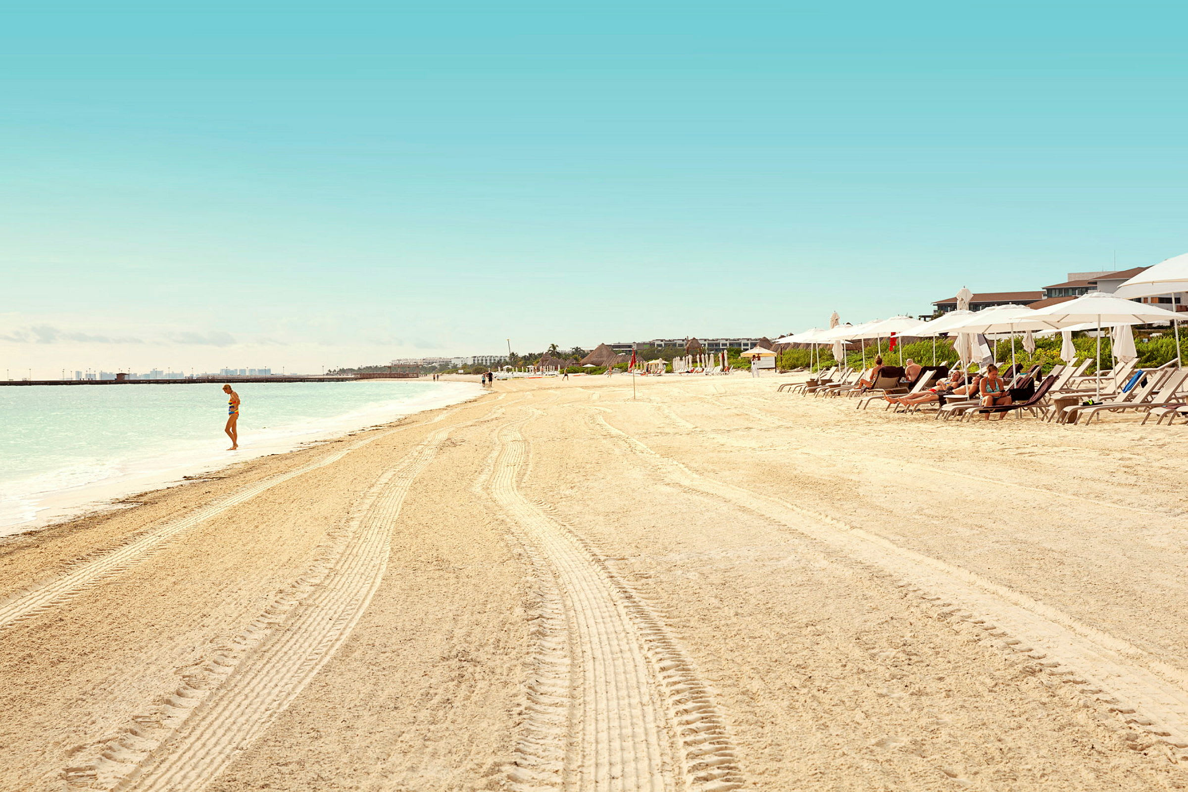 Sandstrand med solsenger og parasoller, folk som nyter solen, og en person ved vannkanten. (AI-generert tekst)