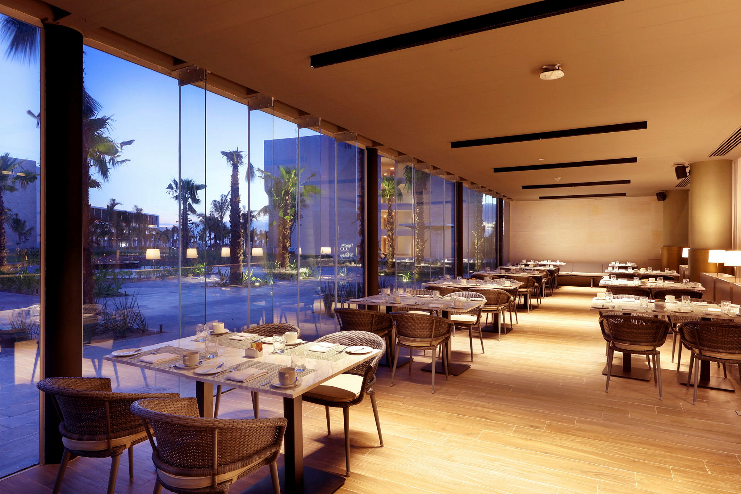 En moderne restaurant med dekkede bord og glassvegger som vender ut mot et utendørsområde med palmer og bygninger. (AI-generert tekst)