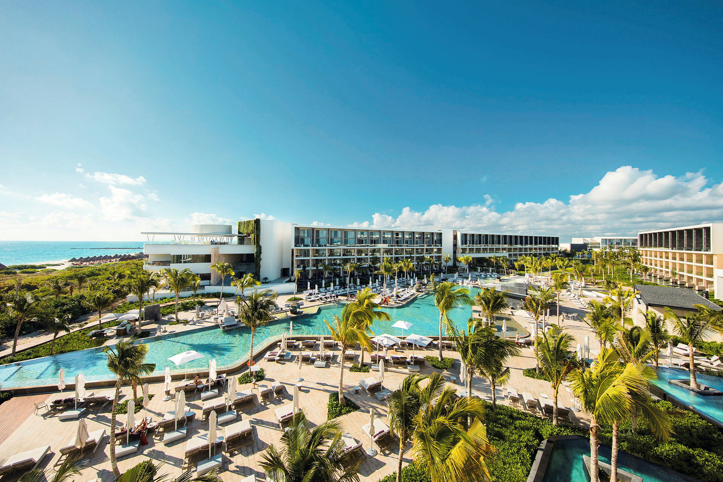 Et moderne hotellkompleks med flere bassenger, frodig vegetasjon og palmer, beliggende ved kysten med utsikt over havet under en klar blå himmel. (AI-generert tekst)