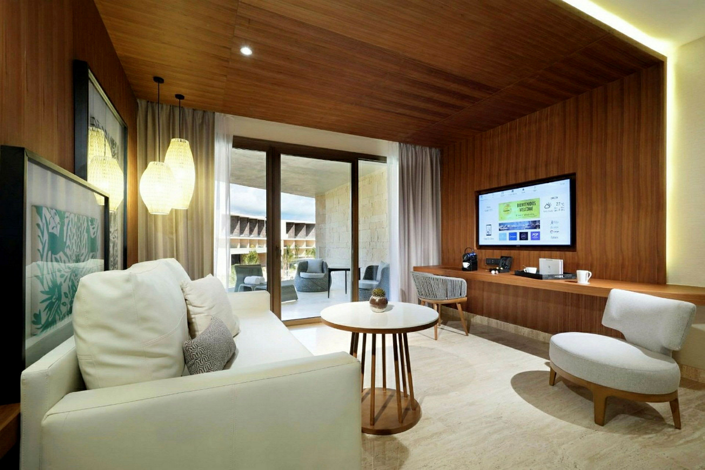 Et moderne hotellrom med tregulv og trepaneler, sofa, et kaffebord og en veggmontert TV. Store glassdører leder ut til en møblert balkong. (AI-generert tekst)