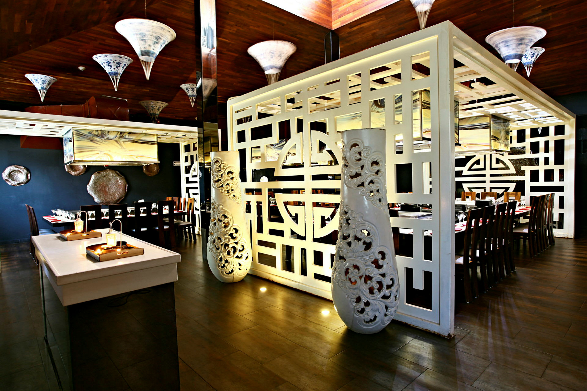 En innendørs restaurant med et elegant innredet område med trebord og stoler. Rommet er dekorert med hvite ornamenterte skillevegger og hengende lamper. (AI-generert tekst)