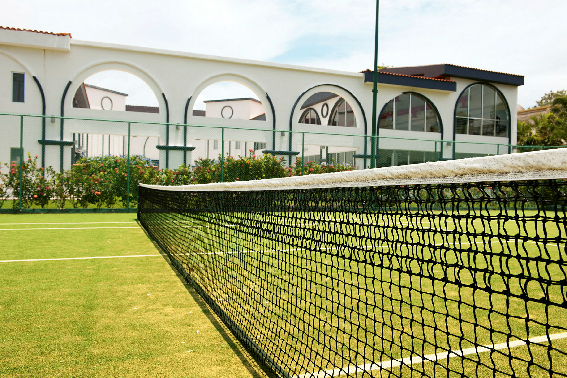 En tennisbane med en grønn overflate og et nett i forgrunnen. I bakgrunnen sees en hvit bygning med buer. (AI-generert tekst)