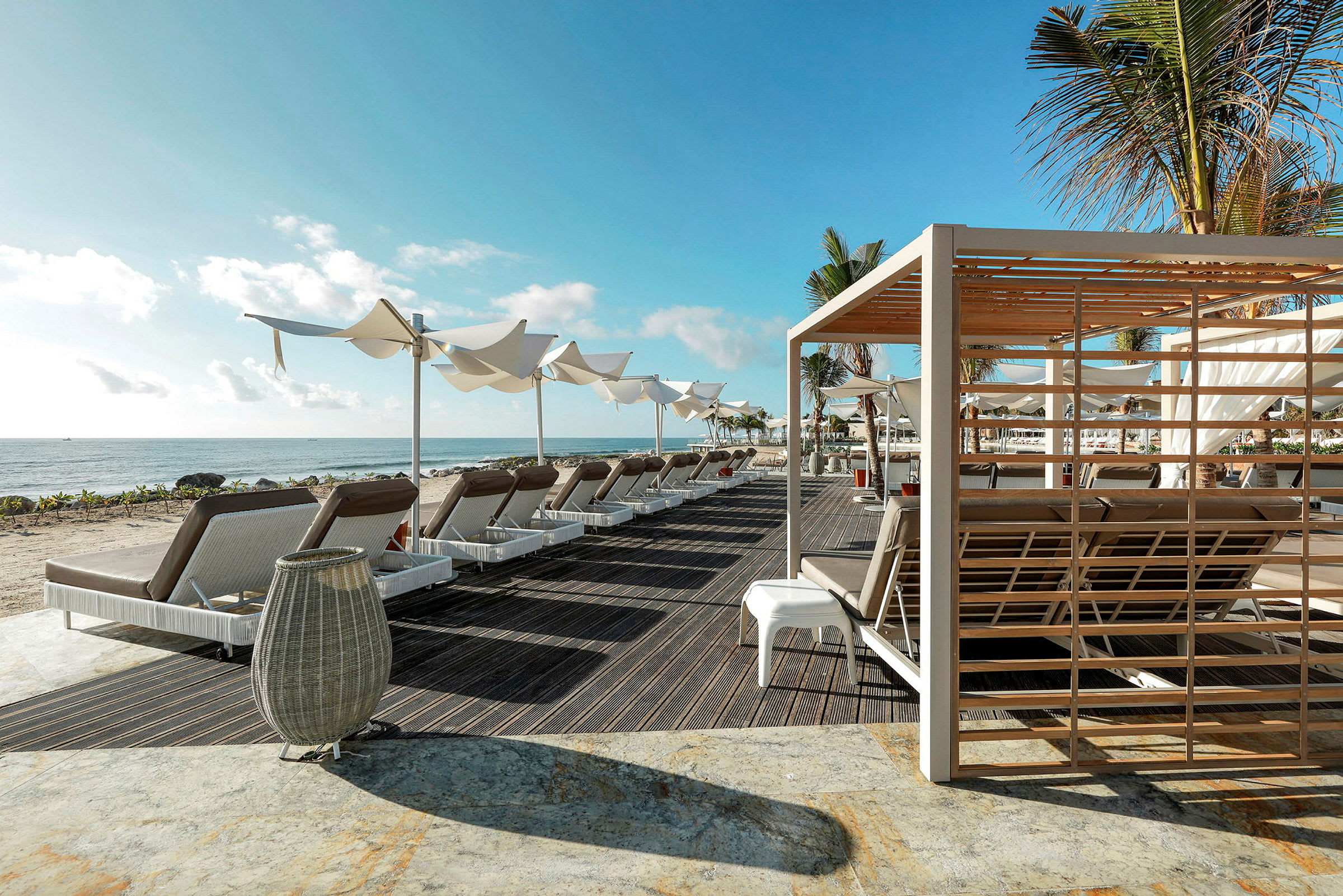 En strand med solsenger, parasoller og en pergola i forgrunnen under en blå himmel. (AI-generert tekst)