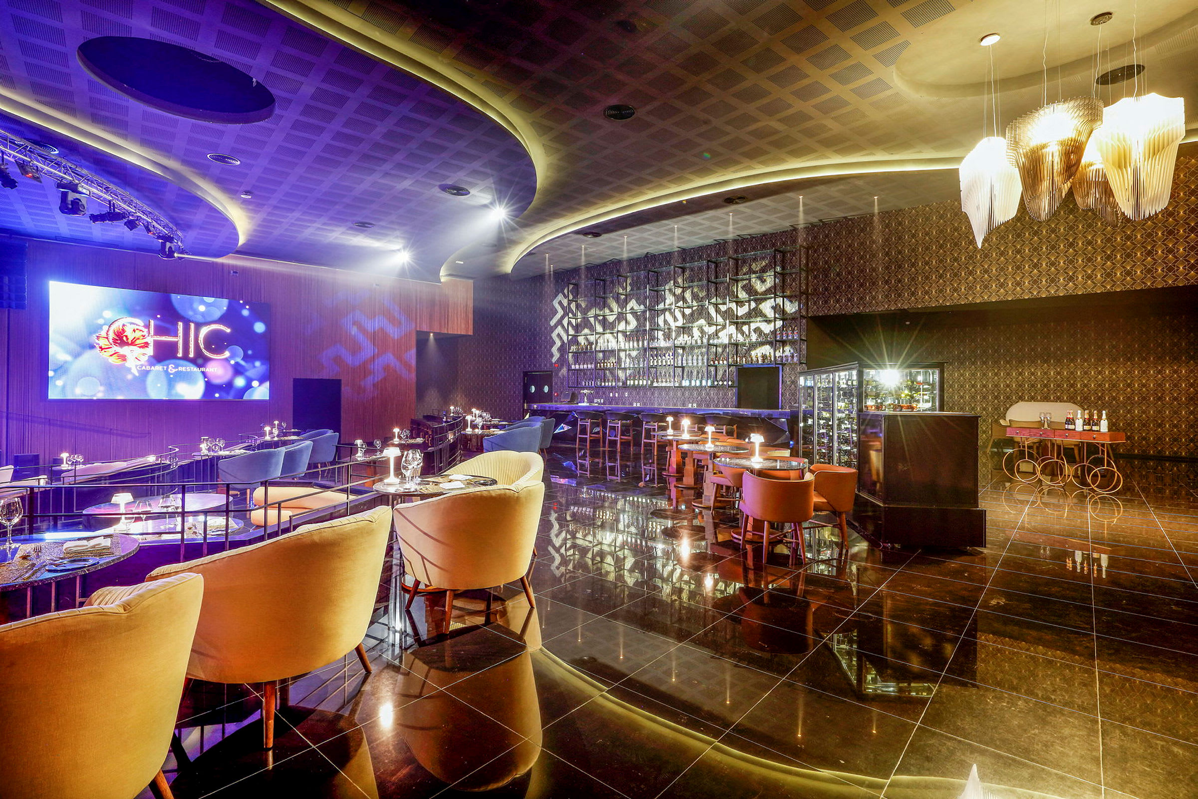 Et elegant innredet bar- og restaurantområde med stemningsfull belysning, luksuriøse sitteplasser og en stor LED-skjerm på veggen. (AI-generert tekst)