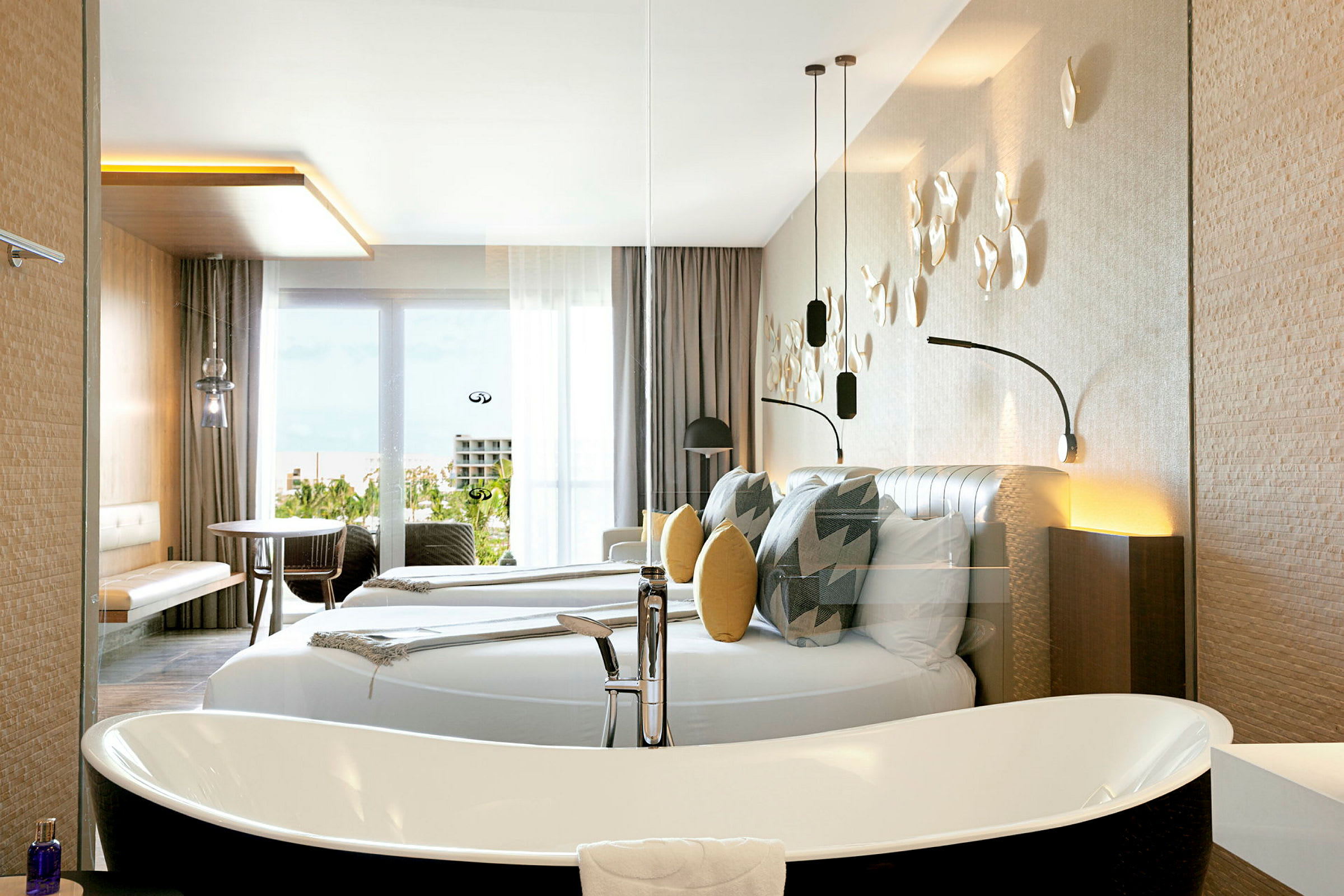 Et moderne hotellrom med en stor dobbeltseng, dekorative puter og veggdekorasjoner. Det finnes et frittstående badekar og møbler som bord og stoler nær en glassdør med utsikt. (AI-generert tekst)