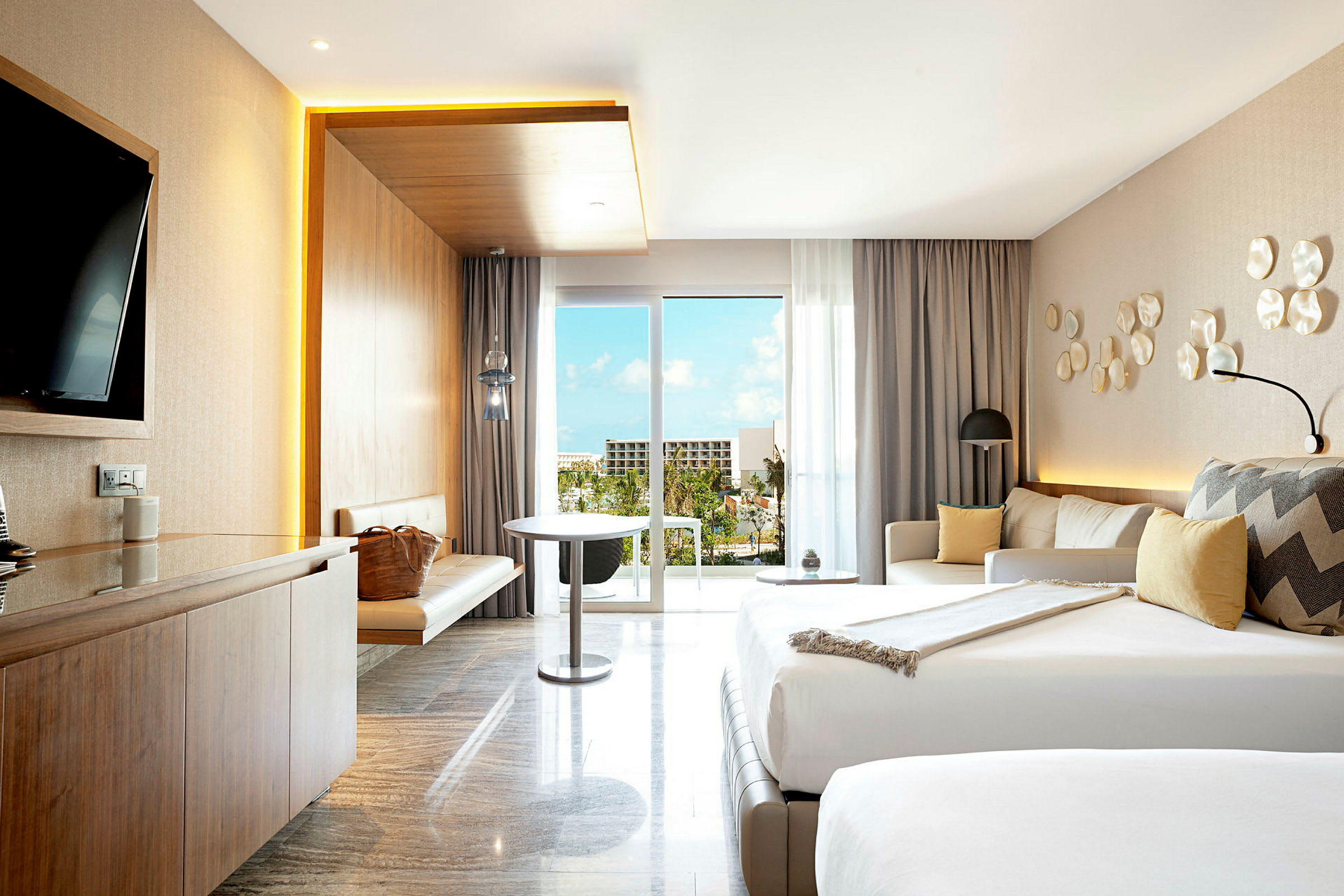 Et moderne hotellrom med dobbeltseng, sofagruppe, bord og TV. Lyse farger og en balkong som vender mot natur og bygninger. (AI-generert tekst)