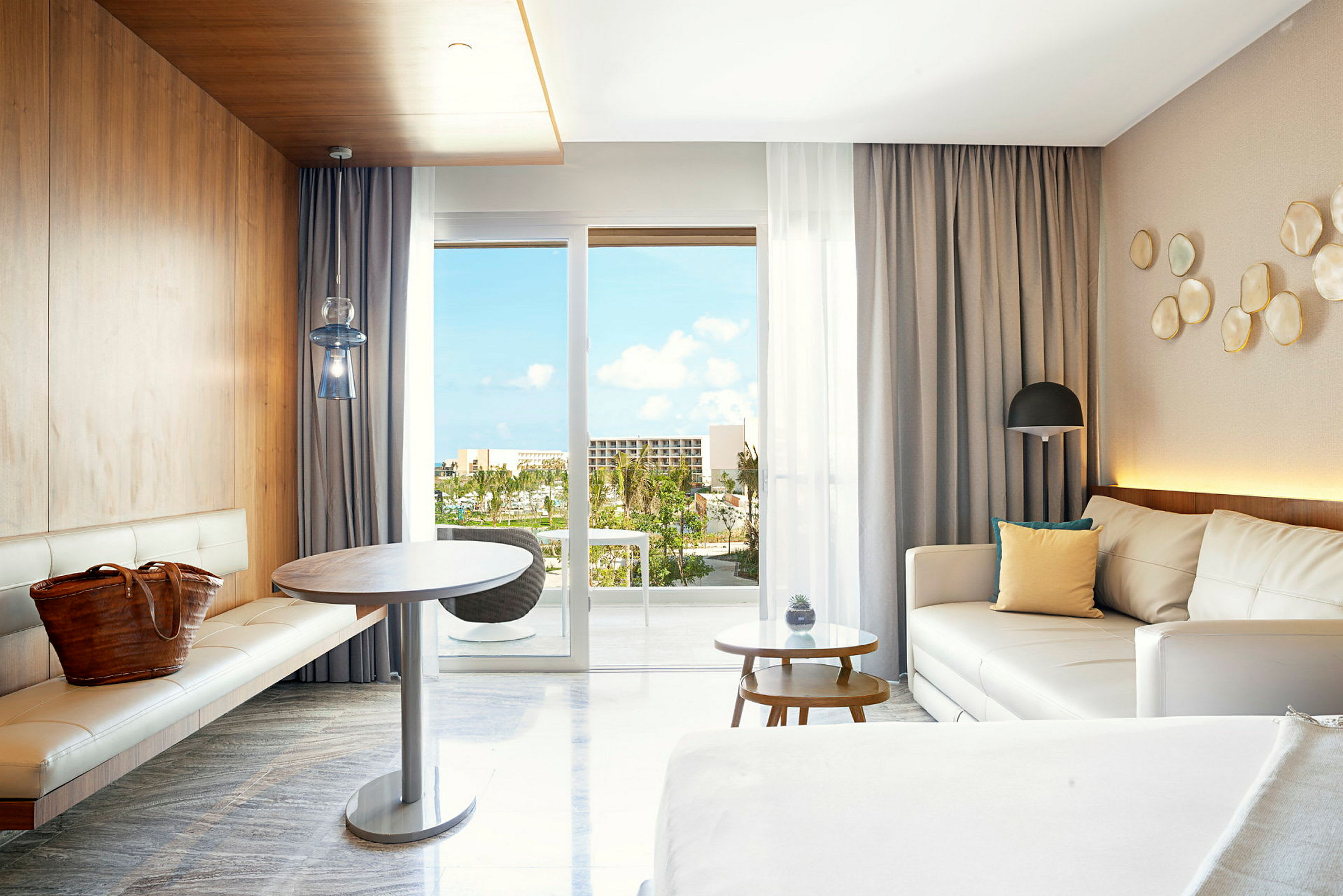 Et moderne hotellrom med hvite møbler og trepanel. Utsikt fra panoramavinduer på balkongen med tropiske planter og blå himmel synlig. (AI-generert tekst)