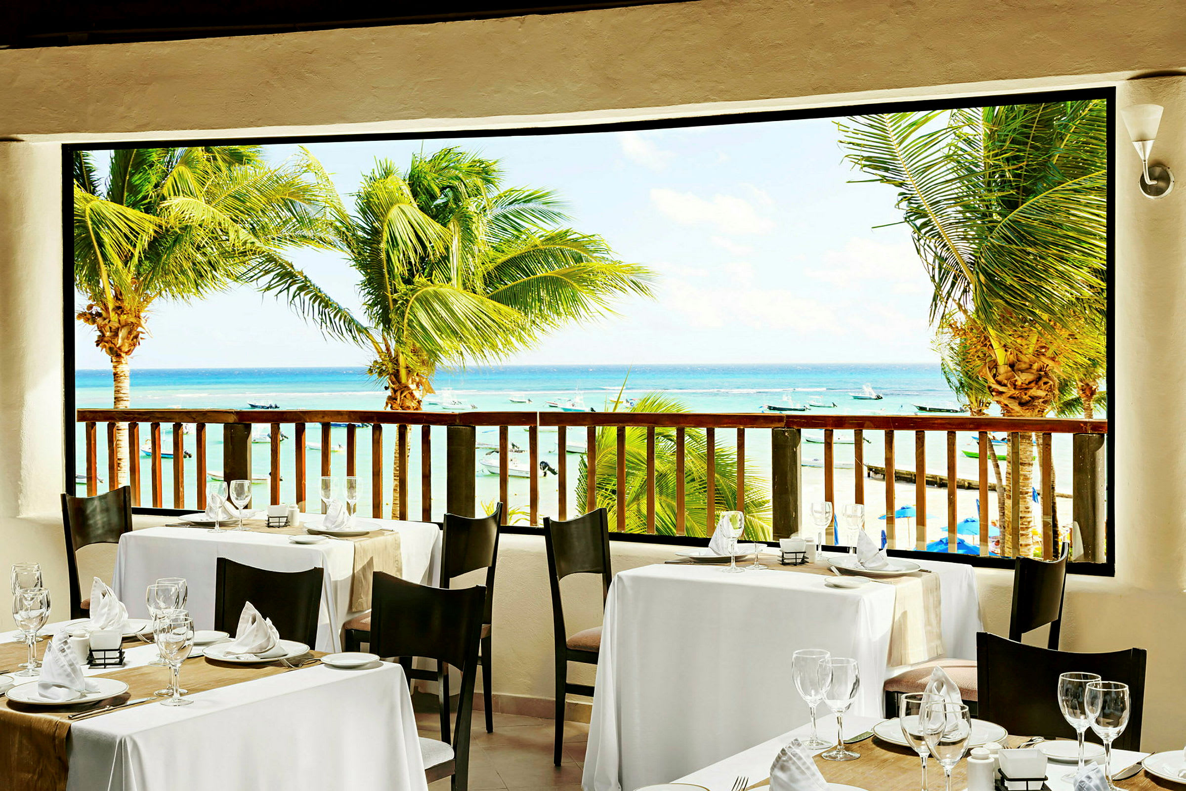 Utsikt fra en restaurant med dekkede bord mot stranden og havet, omgitt av palmer. (AI-generert tekst)