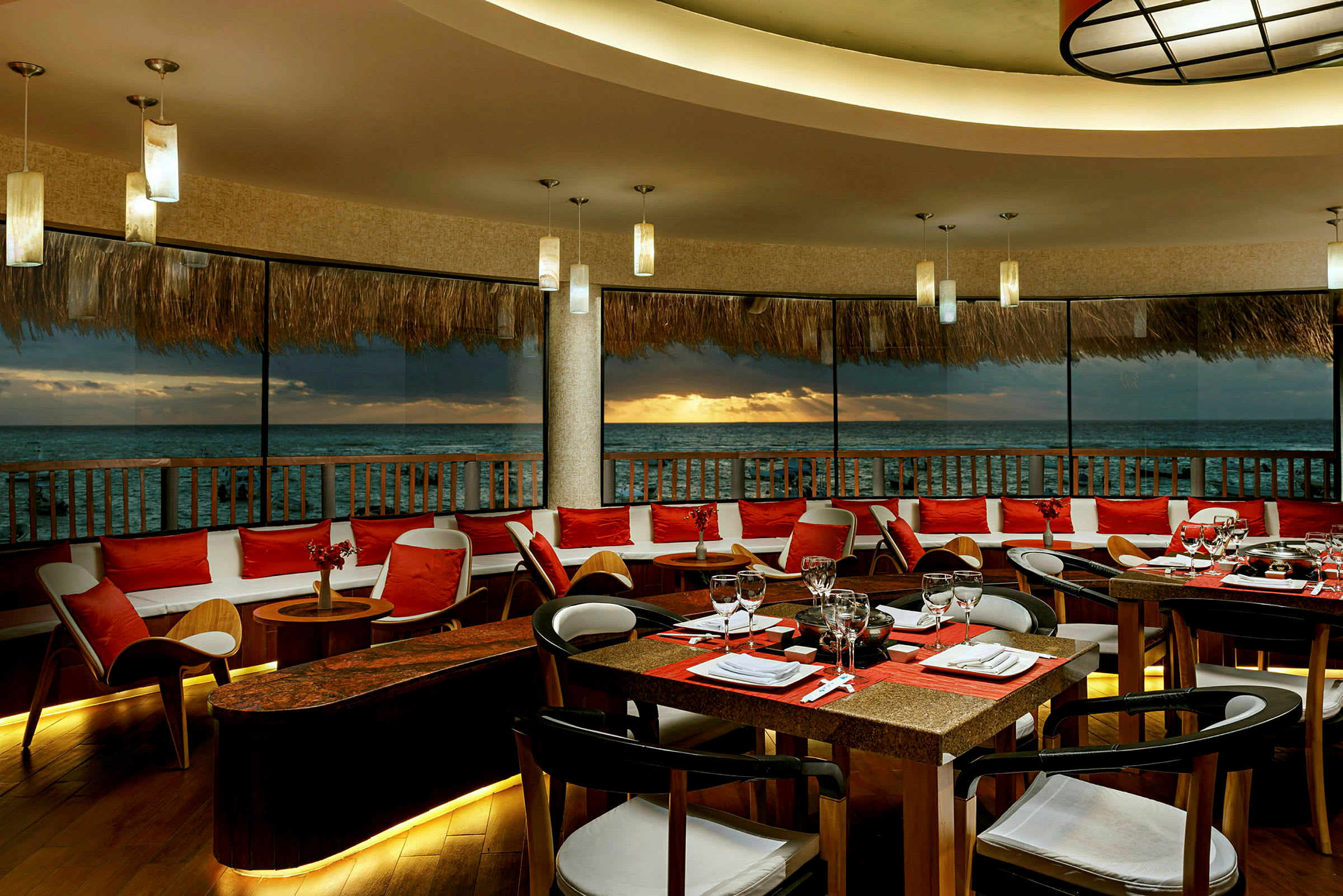 En restaurant med bord og stoler plassert ved et stort vindu med utsikt over havet og solnedgangen. (AI-generert tekst)