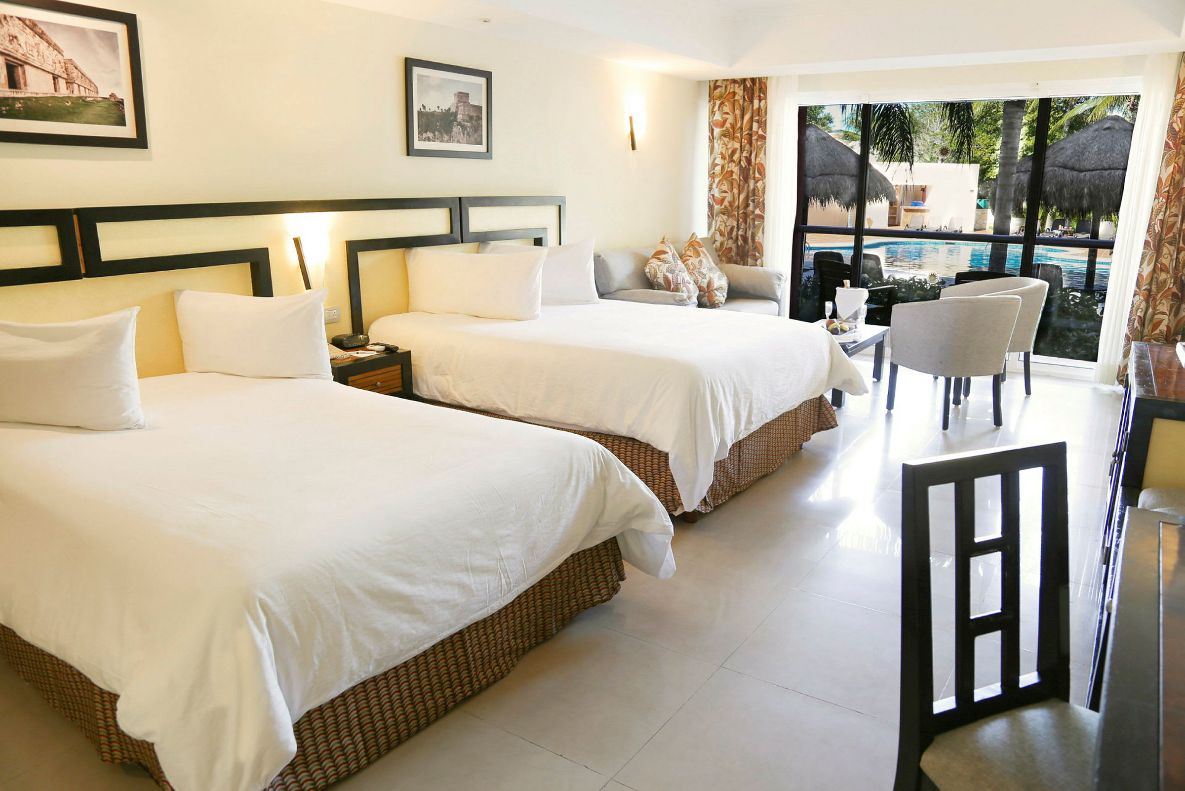 Bilde av et hotellrom med to store senger, sitteområde og et bord foran store vinduer som vender mot et basseng. (AI-generert tekst)