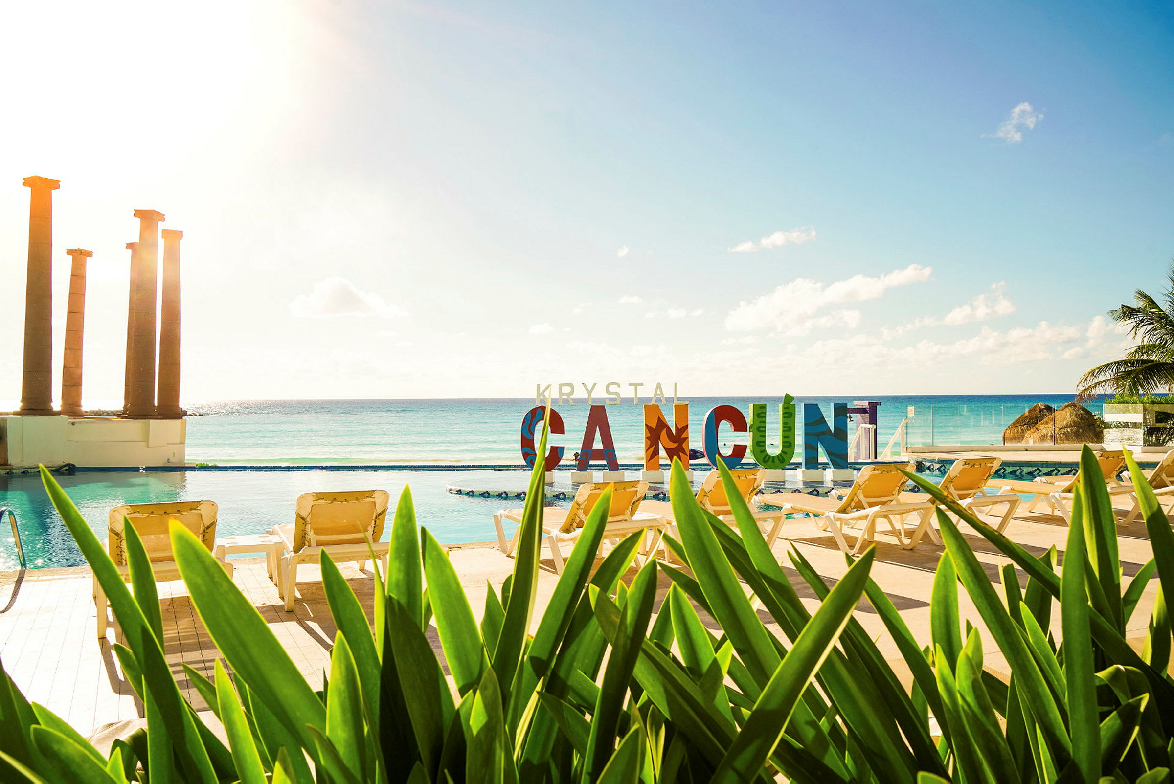 Utsikt over et basseng med solsenger og vegetasjon i forgrunnen. I bakgrunnen ses havet og fargerike bokstaver som staver "CANCUN". (AI-generert tekst)
