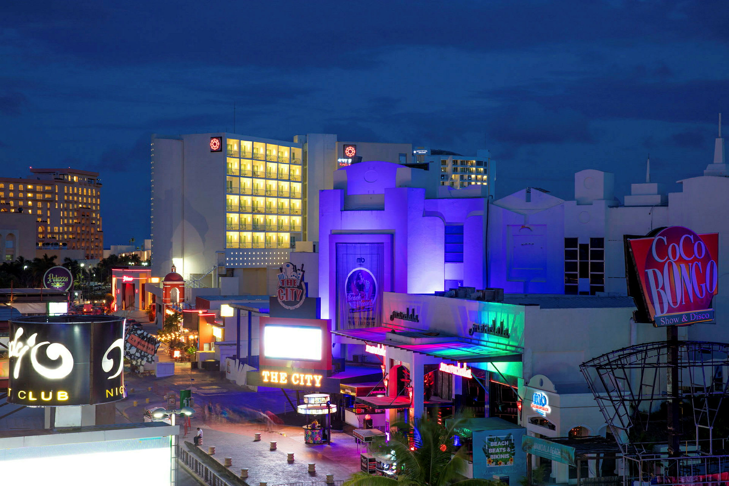 Bilde av et nattklubbområde med fargerike bygninger og skilt, inkludert et tydelig skilt for Coco Bongo. Bakgrunnen viser hotell med opplyste vinduer. (AI-generert tekst)