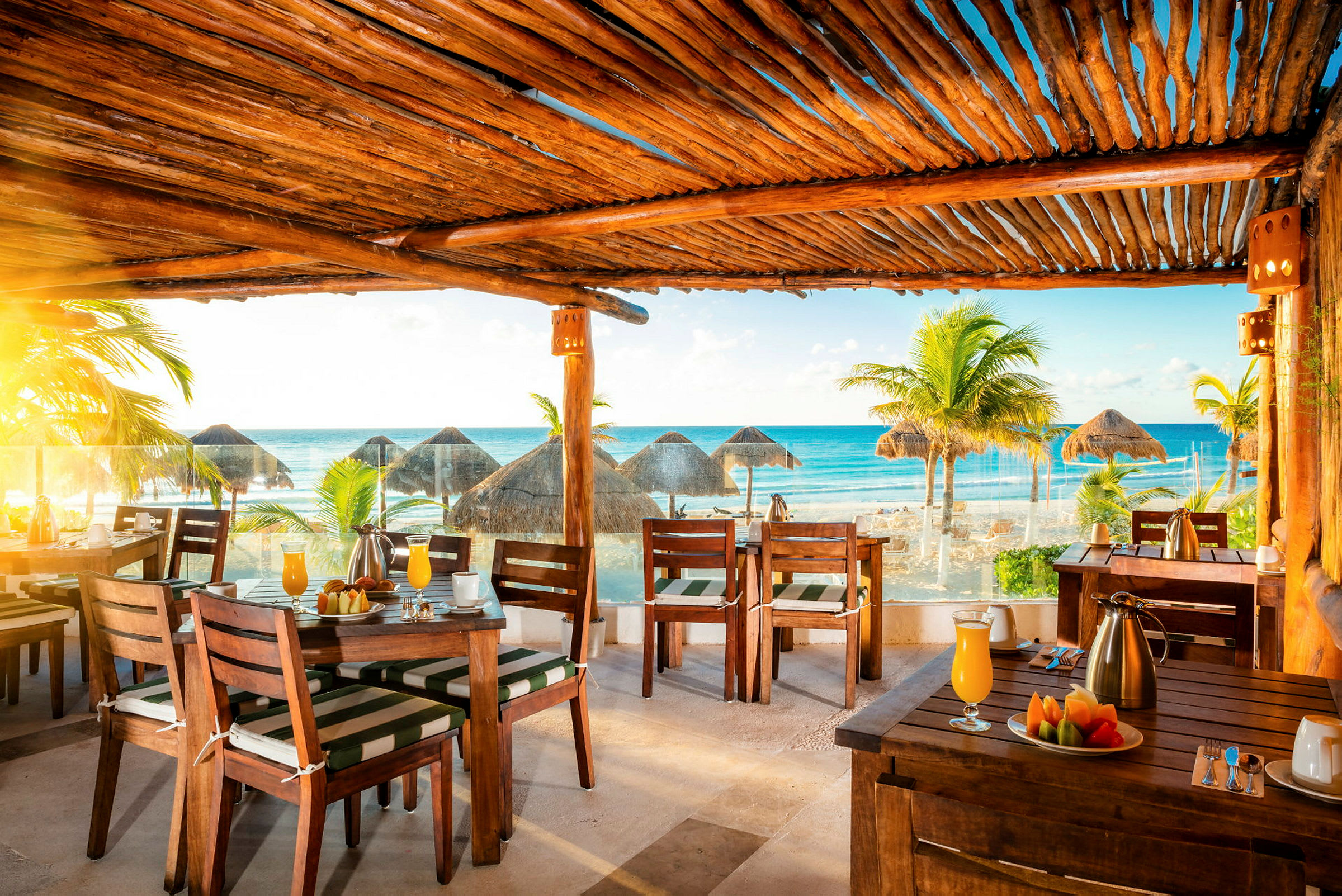 En restaurant med utsikt over stranden og havet. I forgrunnen sees dekkede bord og trestoler. I bakgrunnen er det palmer og stråtak på stranden. (AI-generert tekst)