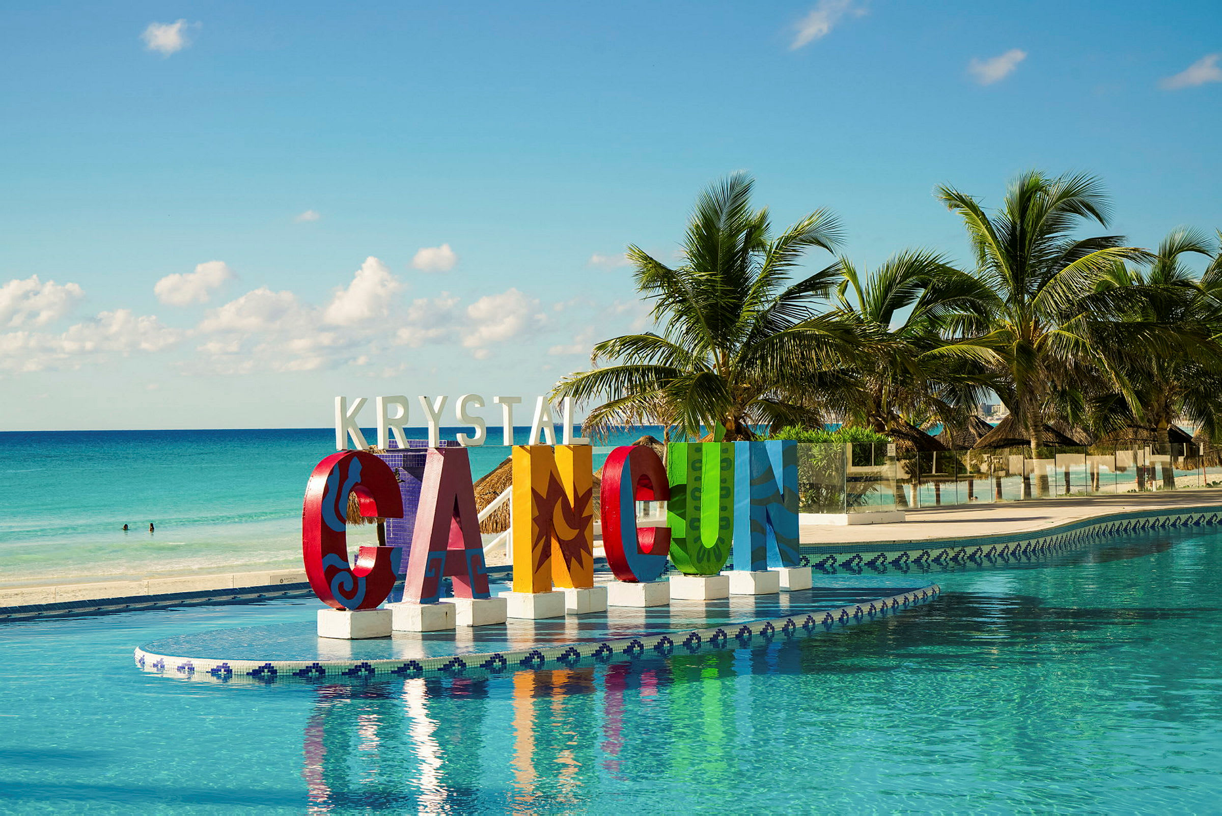 Store fargerike bokstaver som danner teksten 'Cancun' ved et basseng. Palmer og hav i bakgrunnen. (AI-generert tekst)