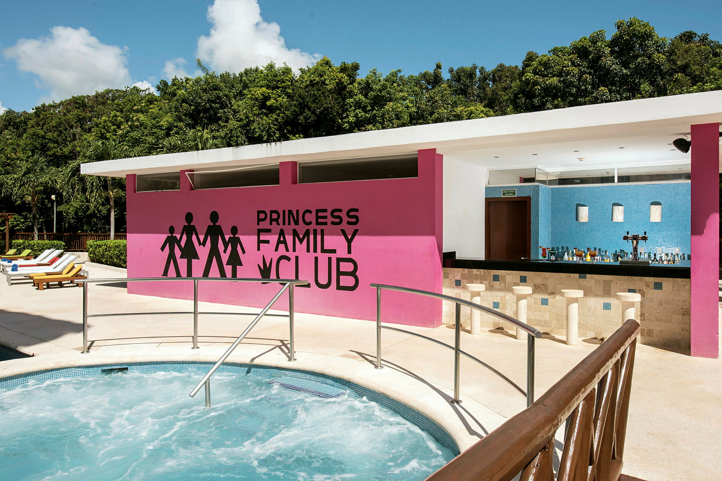 Et utendørsbasseng med et boblebad i forgrunnen, en rosa vegg med teksten 'Princess Family Club' og en bar under et tak. Det er omgitt av grøntområder og solsenger på siden. (AI-generert tekst)