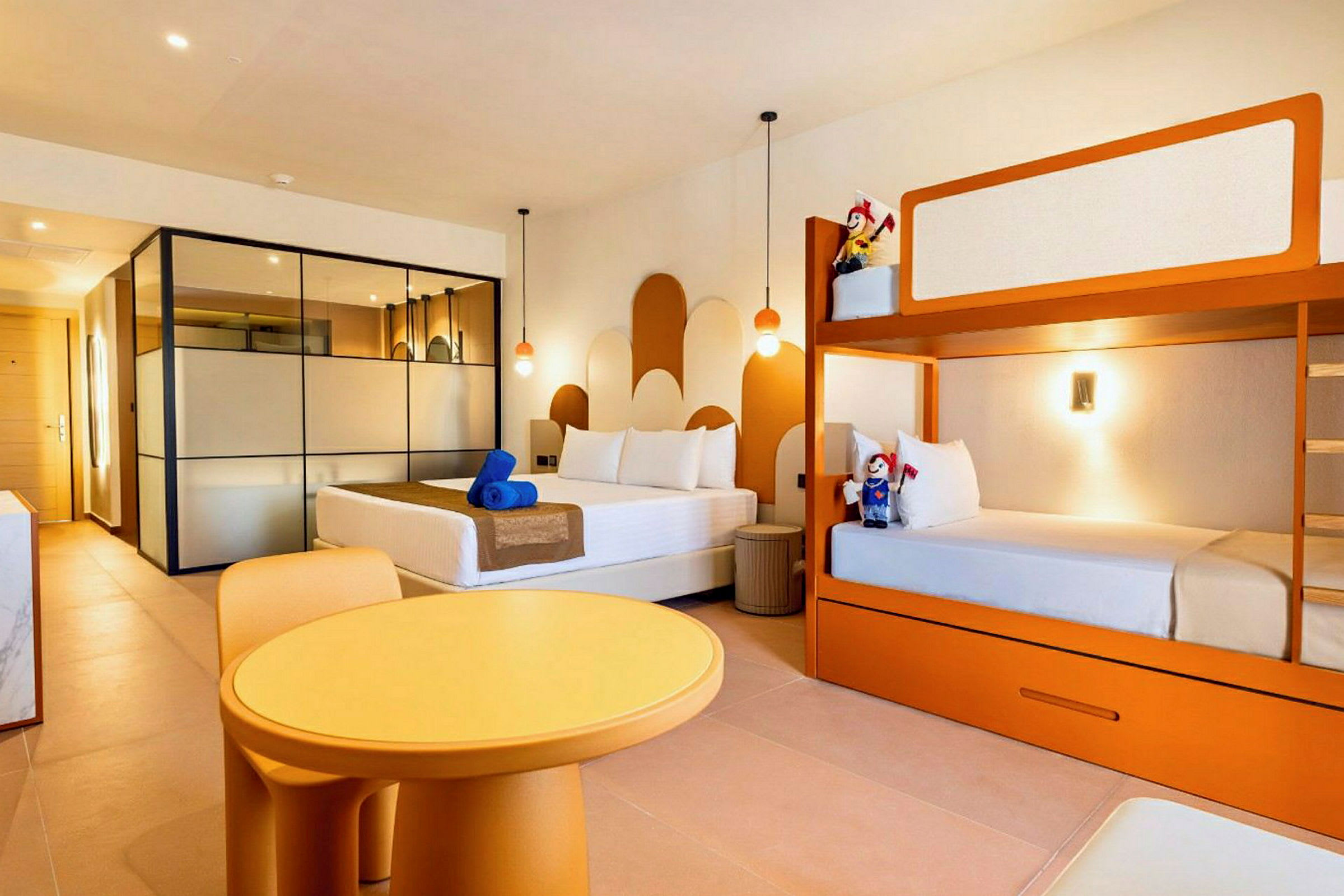 Et lyst hotellrom med en dobbeltseng, køyeseng og et rundt bord med stoler. (AI-generert tekst)