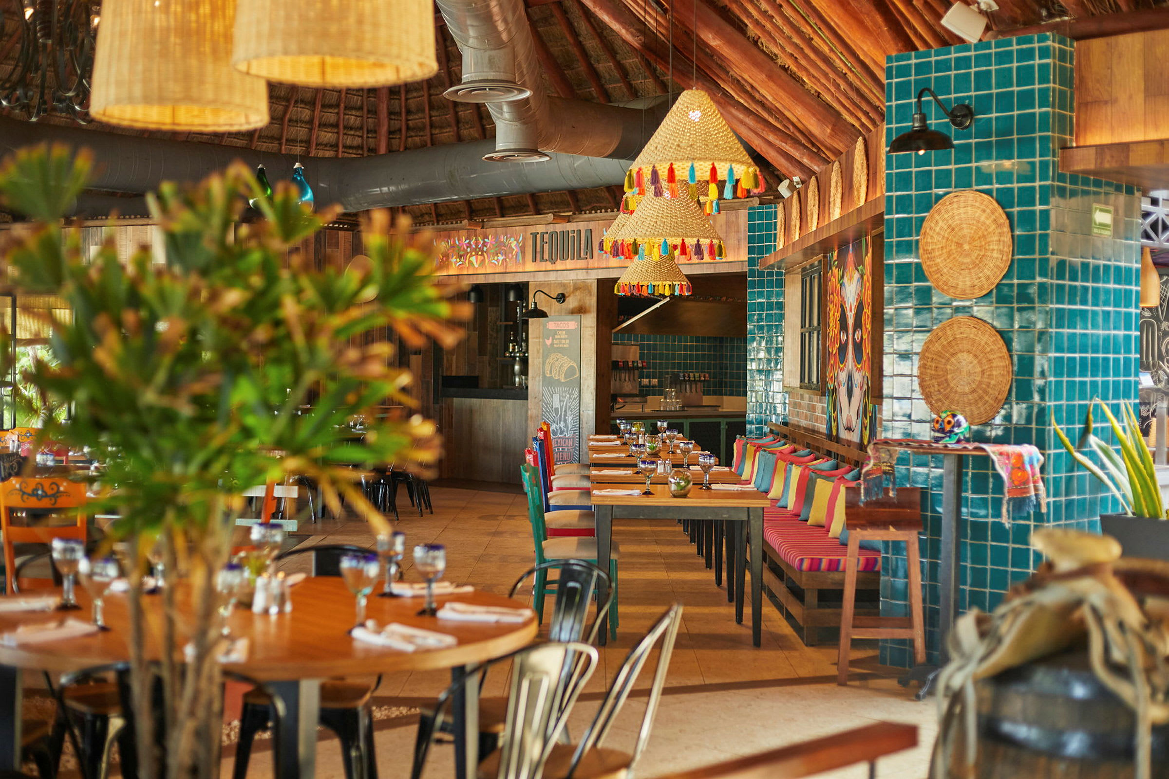 Et fargerikt restaurantinteriør med bord og stoler, dekorasjonene inkluderer puter, grønne planter og hengende lamper. (AI-generert tekst)