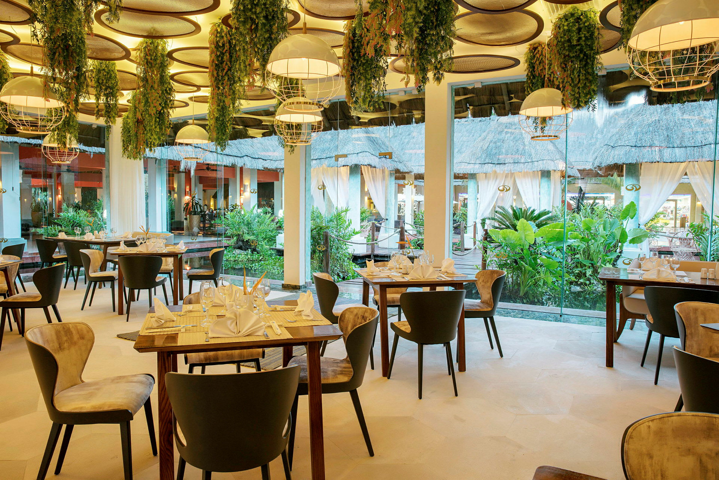 Interiør av en elegant restaurant med dekorerte bord og stoler, hengende lamper og planter, samt store vinduer med utsikt mot en frodig hage. (AI-generert tekst)